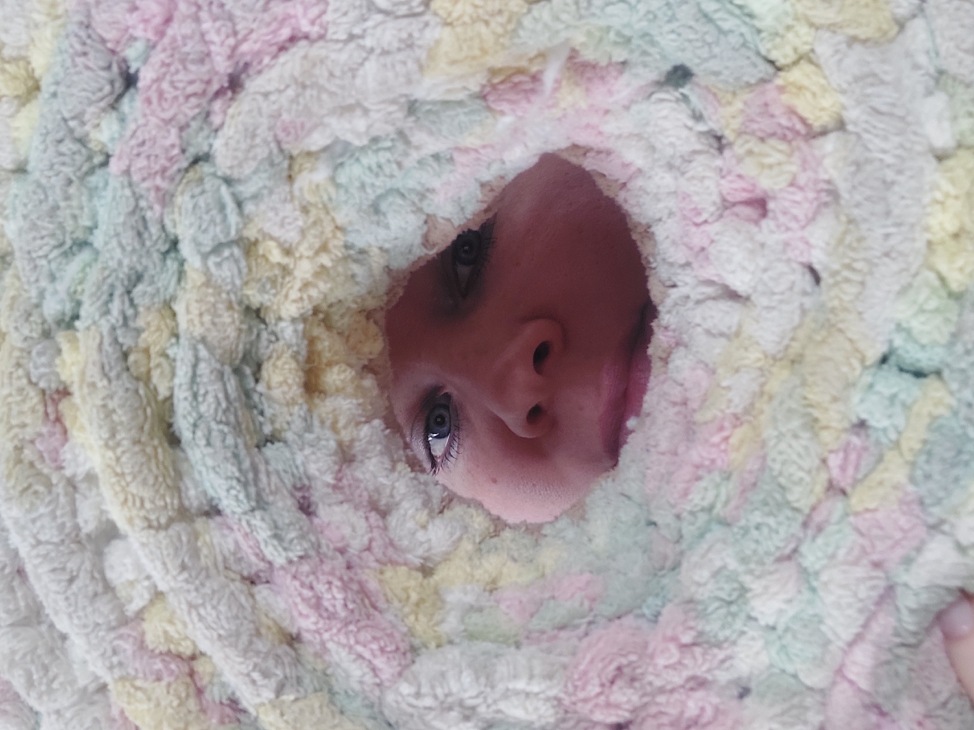 A person's face is visible through a hole in a pastel-colored, looped yarn blanket. The face, featuring a nose, eyes, and a slightly open mouth, is positioned centrally within the circular opening. The blanket, composed of light pink, yellow, green, and white yarns, surrounds the face and creates a textured, tunnel-like effect. The image appears to be taken from a low angle, looking up through the blanket towards the face. It is not possible to determine the location as Chaikovsky, Russia from the image alone.