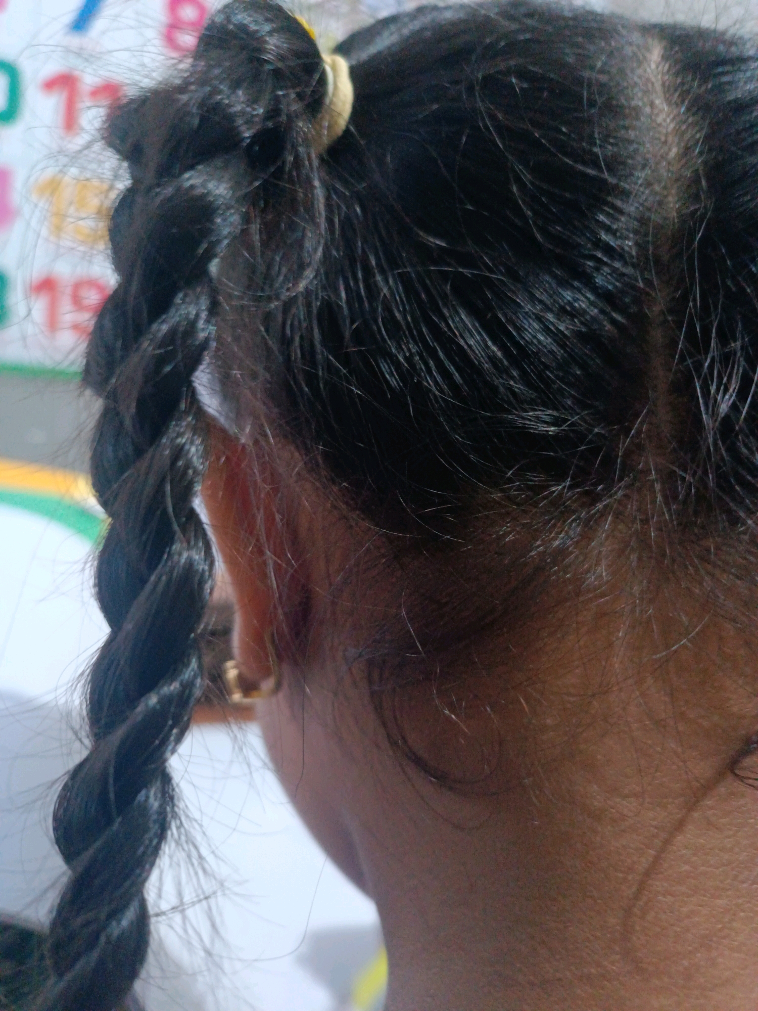 The image shows a close-up, rear view of a young girl's head and neck. Her dark hair is braided into a thick, visible plait on the left side of the frame, secured with a light-colored hair tie. Some fine strands of hair are loose around her head. She is wearing a small, gold-colored earring in her visible left ear. The background is blurred but appears to be a surface with colorful numbers and markings, possibly a chart or educational material. The lighting suggests an indoor setting. The scene is likely from a home environment in Tabanan, Indonesia.