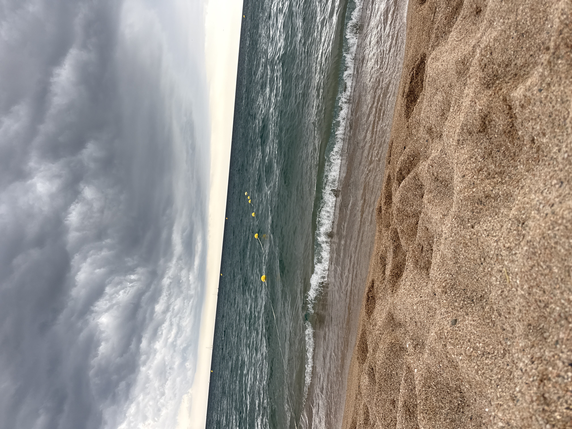 The image depicts a beach scene in Blanes, Spain. The foreground is dominated by wet sand with footprints and ripples, suggesting the tide has recently receded. The middle ground features the turquoise waters of the Mediterranean Sea, with small waves lapping at the shore. Several yellow buoys are visible in the water, marking a designated swimming area or a boundary. In the background, the horizon is marked by a pale, overcast sky filled with dramatic, dark grey storm clouds. The overall impression is one of an impending storm, with the sea appearing choppy and the sky dark and heavy. The scene is devoid of any people or boats.