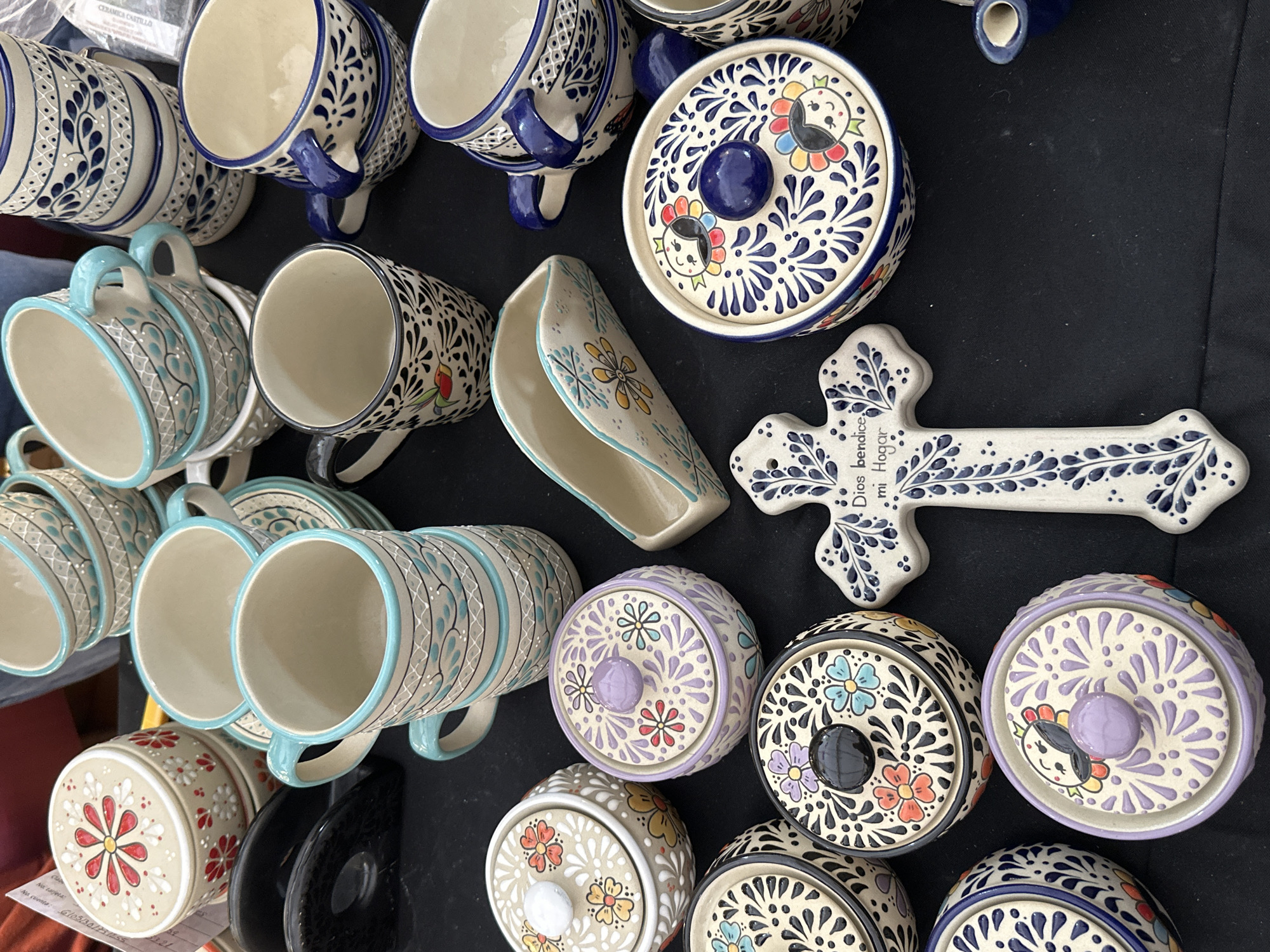 An array of handcrafted ceramic pottery is displayed on a flat black surface, likely a market stall in Mexico City, Mexico. The collection includes various mugs, lidded containers, a napkin holder, and a decorative cross, all featuring distinct hand-painted patterns.
In the upper-left, three cream-colored cylindrical lidded containers are stacked, each adorned with red and yellow stylized floral designs. Adjacent to these is a black circular object. In the upper-center, a stack of four mugs features light blue rims and handles with light blue and white intricate patterns. To their right, another stack of four mugs exhibits dark blue rims and handles with similar patterns in dark blue and white. A third stack of three dark blue-rimmed mugs is visible further right.
In the mid-left, a mug with a black handle is decorated with black and white patterns and a small, colorful bird illustration. Below this, three round lidded containers with dark blue and white patterns are partially visible. A light-colored round lidded container in the lower-left corner displays black, orange, and blue floral patterns.
Centrally, a light purple round lidded container features light purple swirls and multi-colored flowers. Below it, another round lidded container with a dark lid shows black, orange, and blue floral motifs. A white ceramic cross, positioned in the lower-center, is decorated with dark blue foliage patterns and inscribed with "Dios bendice mi Hogar" (God bless my Home).
To the mid-right, a cream-colored ceramic napkin holder with light blue trim and yellow floral accents is visible. In the lower-right, a dark blue round lidded container showcases stylized faces within colorful floral borders and dark blue patterns. A small dark blue creamer or pitcher is partially visible in the bottom right corner.
The items are arranged in a dense, organized fashion, characteristic of a retail display. The background is blurred, indicating an out-of-focus environment consistent with a public setting such as an outdoor or indoor market.