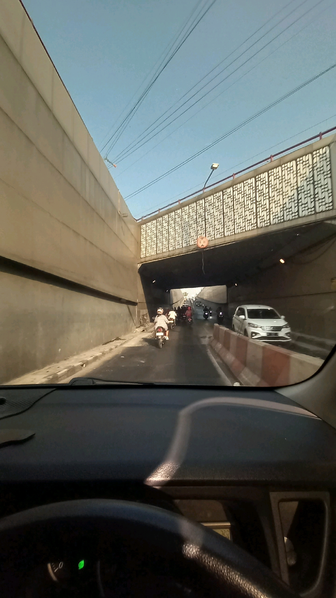 A first-person perspective shot captures traffic moving through an underpass in Purbayan, Indonesia. The road surface is wet, suggesting recent rain or a cleaning operation. A white car is visible in the right lane, and several motorcycles with riders are navigating the underpass. Overhead, power lines crisscross the bright blue sky, with a decorative bridge structure spanning the entrance to the underpass. The scene is framed by the interior of a vehicle, including the dashboard and steering wheel.