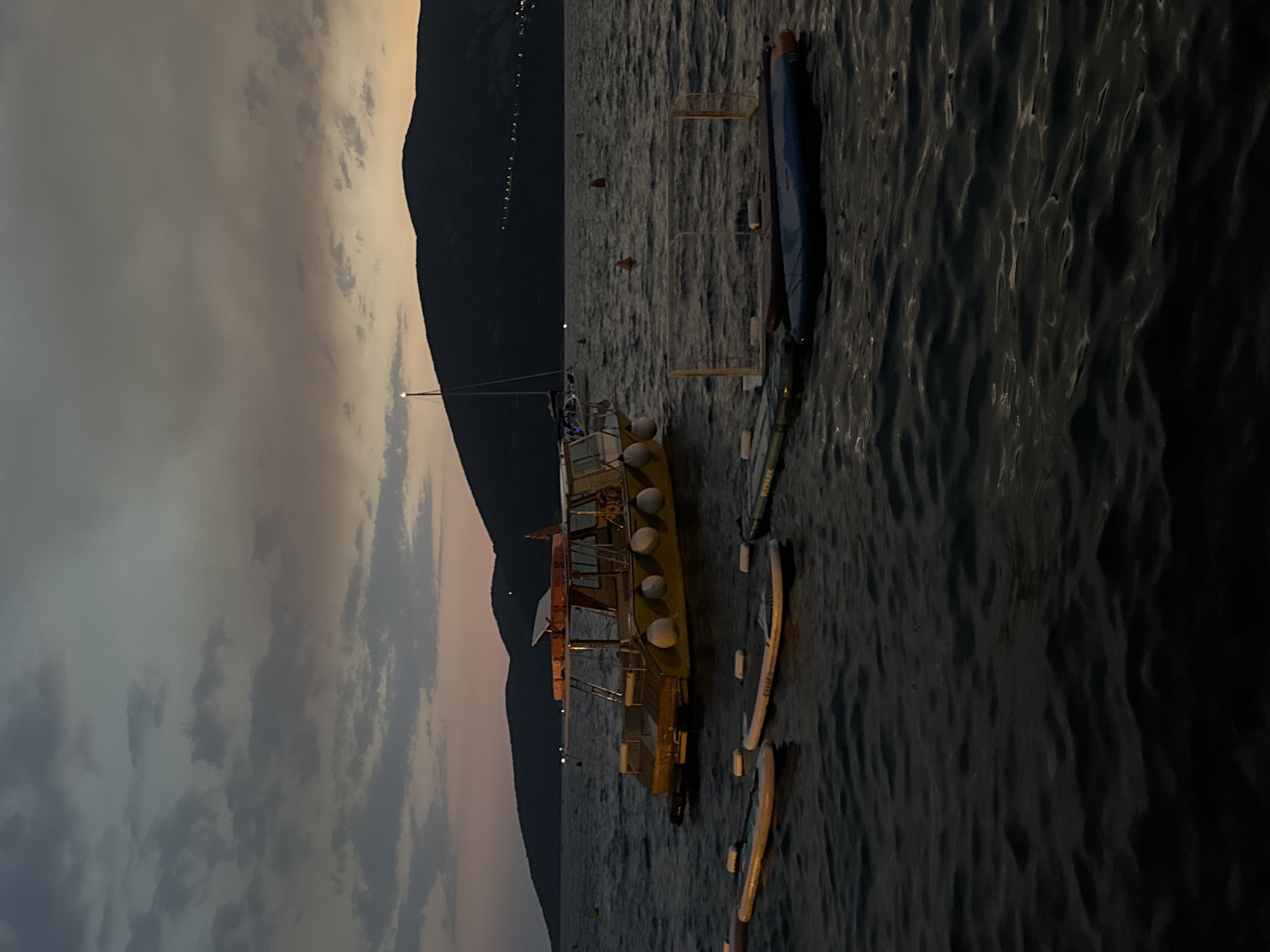 The image depicts a serene twilight scene at sea, likely near a coastal city, though the exact location remains unknown. The sky is a canvas of muted grays and oranges, with scattered clouds hinting at the fading light. In the foreground, the dark, rippling water reflects the dim sky. Several inflatable paddleboards are visible, some tethered to buoys, suggesting a recreational area. Further out, a yellow boat with a covered top is anchored, its sides dotted with white fenders. A sailboat with a tall mast stands in the background, silhouetted against the dusky sky. Beyond the water, a dark, undulating silhouette of hills or mountains stretches across the horizon, with faint lights twinkling on their slopes, indicating a distant settlement. The overall atmosphere is one of calm and tranquility, a peaceful end to a day.