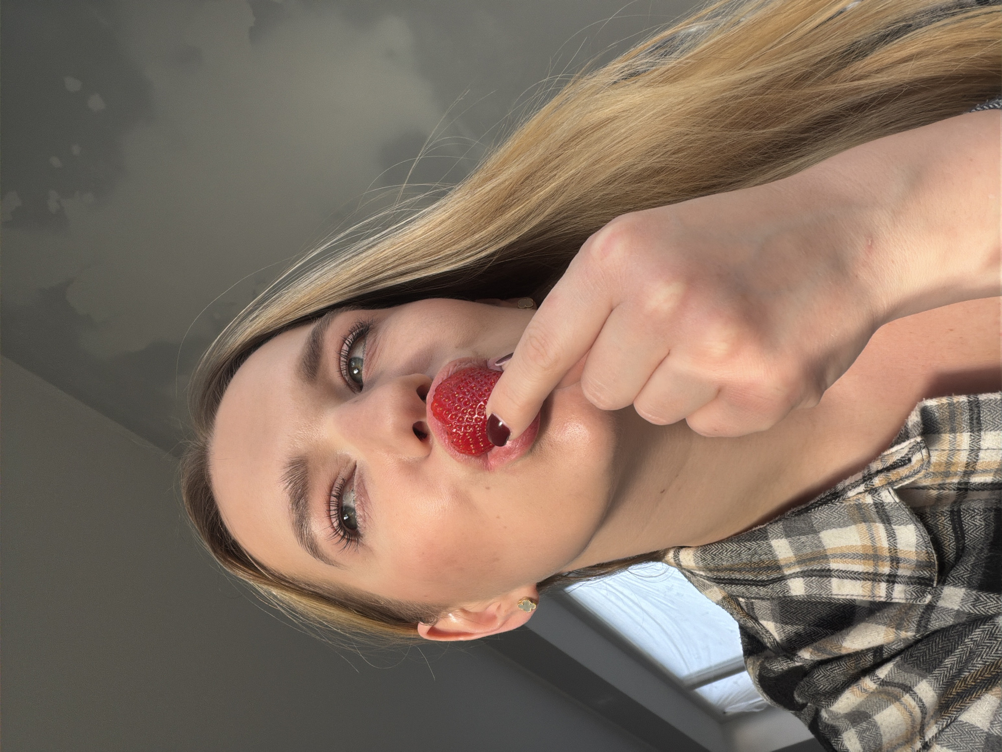 A young woman with blonde hair holds a strawberry to her lips. The background appears to be an overcast sky. She is wearing a plaid shirt. The photo was taken from a low angle, looking up. It is not possible to verify that this image was taken in Dreiliņi, Latvia.