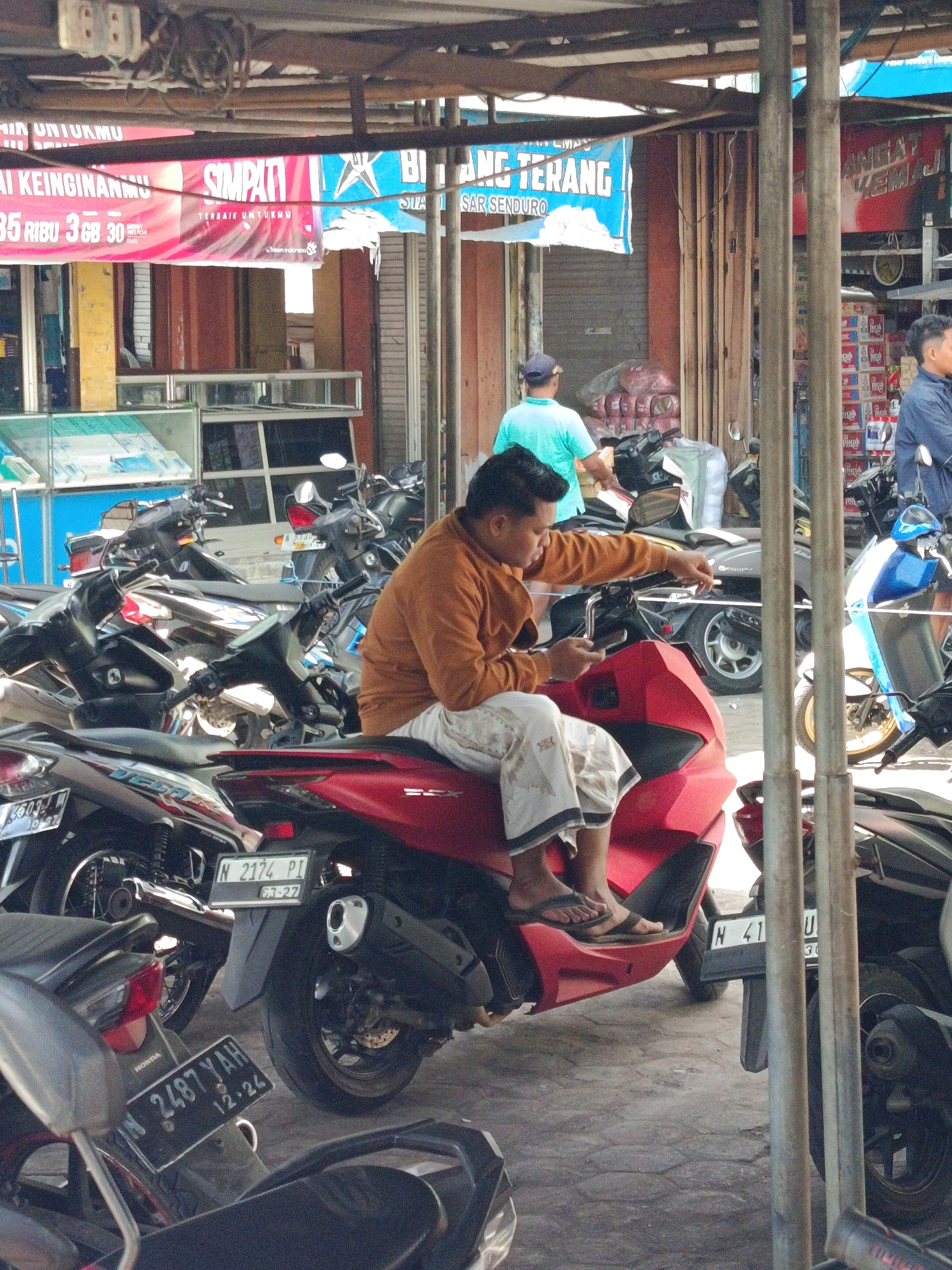 The image captures a street scene in Senduro, Indonesia, with a focus on motorcycles parked in what appears to be an outdoor market or commercial area. In the foreground, a man wearing an orange long-sleeved shirt and white pants with a patterned border is sitting on a red scooter. He is holding a smartphone in his left hand and gesturing with his right hand towards the phone. His feet, clad in black sandals, are resting on the scooter's footrest.
Behind the man, several other scooters and motorcycles are visible. Some are parked neatly, while others are more crowded together. License plates are visible on some of the motorcycles, including one with the plate number "N 2174 PI" and another with "N 2487 YAH". The ground is paved with concrete.
In the background, there are several stalls or shops with various goods displayed. A red banner with white lettering is prominently displayed, partially obscured by a metal support. Text on the banner includes "SIMPATI" and what appears to be promotional information like "TERBAIK UNTUKMU" and "35 RIBU 3 GB 30 HARI". Another banner to the right reads "ANGAT TERANG". A person in a turquoise shirt stands near a stall, and another person in a dark jacket is partially visible on the far right. The overall environment suggests a bustling commercial area.