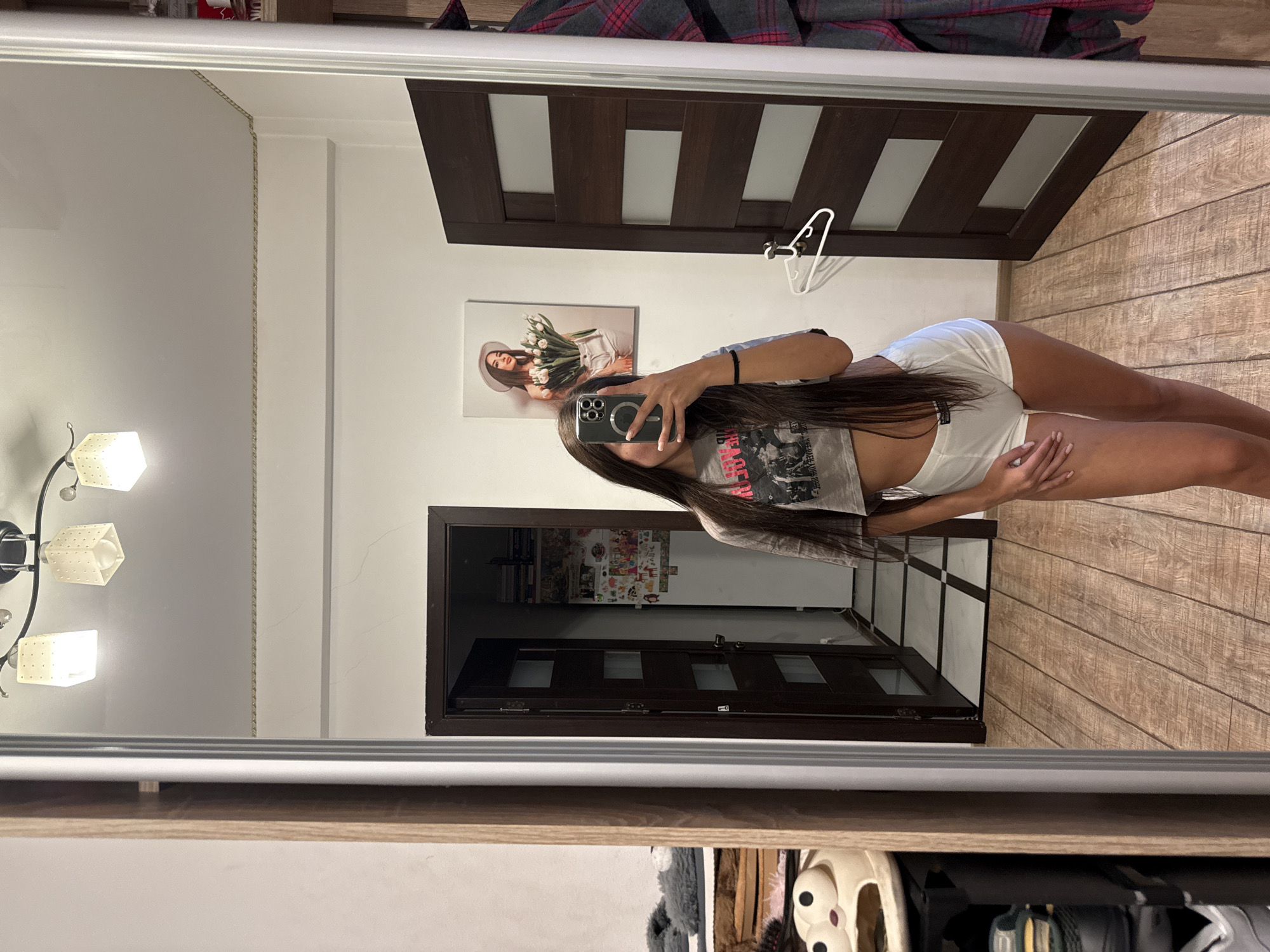 A young woman with long dark hair takes a selfie in a mirror. She is wearing a grey cropped t-shirt and white shorts. The room has wooden floors and two dark wooden doors. On the wall, there is a framed picture of a woman holding tulips. A closet with clothes hanging on the right side of the frame is also visible. The scene appears to be in a residential interior.