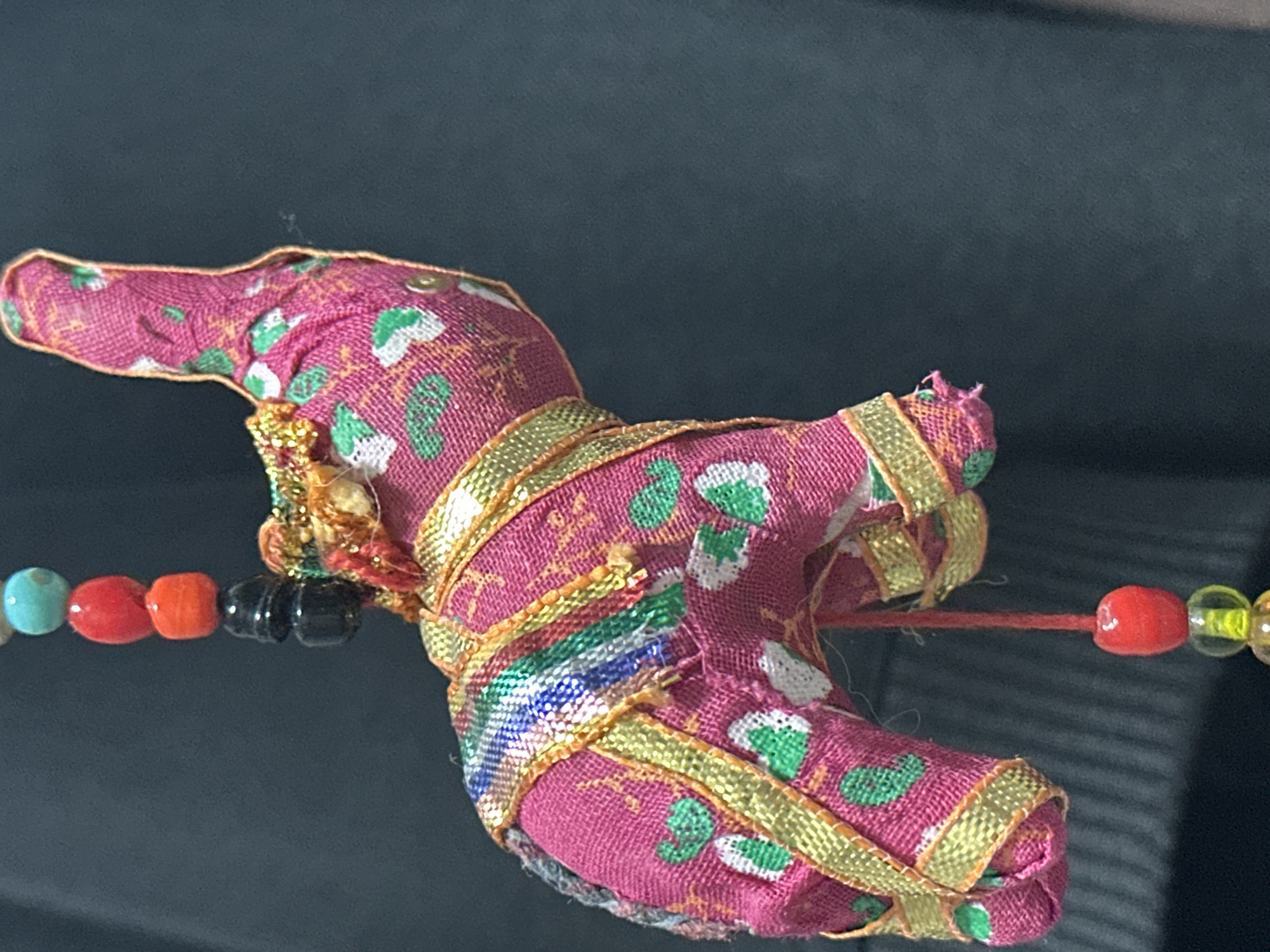 The image is a close-up shot of a fabric elephant ornament, primarily colored pink with green and white floral patterns. It is adorned with gold ribbon accents and a multi-colored beaded string hangs from its underside, featuring beads of red, orange, black, turquoise, and clear yellow. The ornament is suspended against a dark, blurred background, possibly clothing or fabric. The image does not contain any information about the city of Nevinnomyssk, Russia.