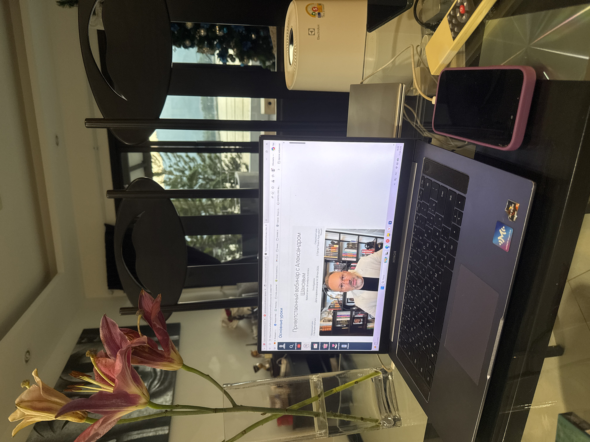 On a glossy black table sits an open Honor laptop, displaying a webpage with a man in a white shirt and glasses on the screen. The text on the page is in Russian. To the right of the laptop is a purple-cased phone charging, a small remote, and an Electrolux device. In front of the laptop, a clear rectangular vase holds lilies. In the background, a black decorative room divider stands near a window, revealing a blurred outdoor scene. The exact location within Baan Chaweng, Thailand, cannot be determined from the image.
