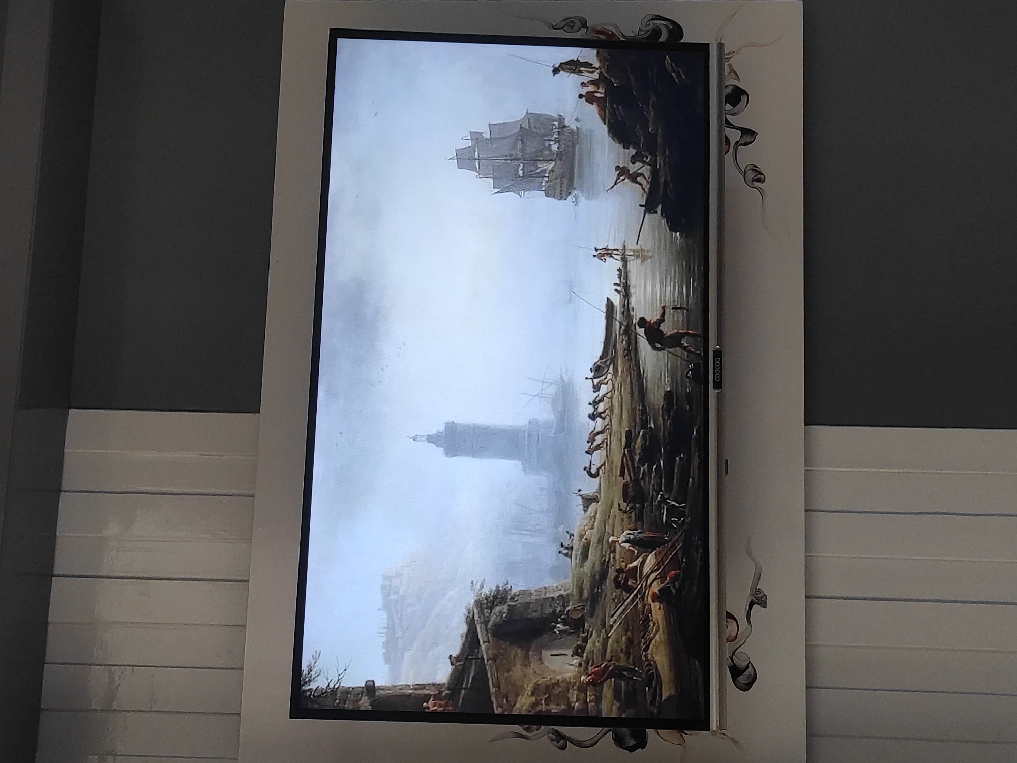 A painting of a harbor scene is displayed on a flat-screen television. The painting depicts a foggy morning with a lighthouse in the background. Several people are shown on the shore, some pulling a boat, others fishing. A large sailing ship is anchored in the harbor. The television is mounted on a wall with a decorative border. There is no indication of the location being Sumedang, Indonesia.