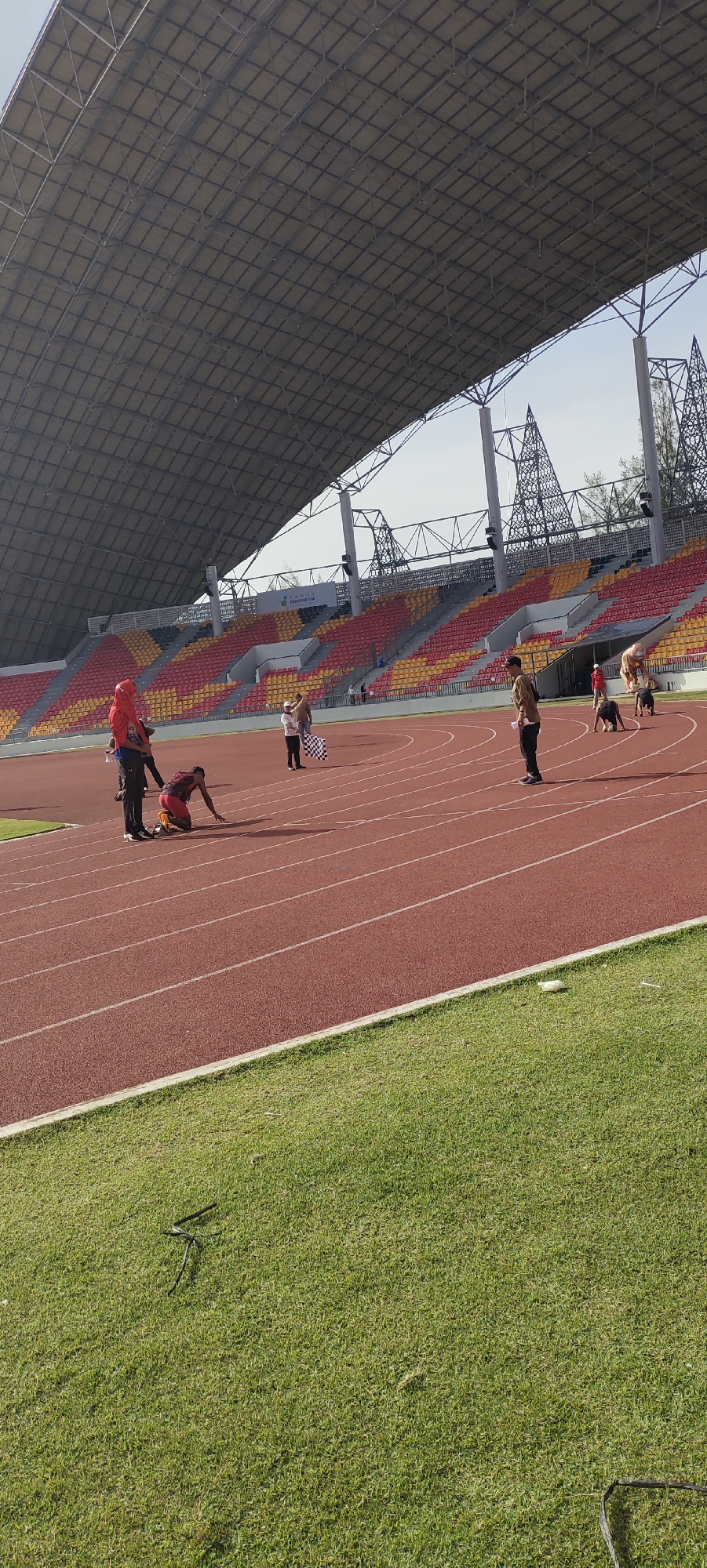 In an outdoor athletic stadium in Banda Aceh, Indonesia, a group of individuals are positioned on a red running track. The stadium features tiered seating in red and yellow, partially covered by a large, gray, metallic roof structure. The track is bordered by a green grass field.
In the foreground, a person in a red hijab and black pants stands on the left side of the track, facing right. To their right, a runner in a red and black racing outfit is on their knees on the track, seemingly at a starting position. Further down the track, another individual stands holding a black and white checkered flag, likely indicating the finish line. Several other people are scattered on the track in various postures, some standing and some appearing to be in a crouched position. The lighting suggests it is daytime, with shadows cast by the individuals on the track. The overall scene depicts a moment of athletic preparation or competition.