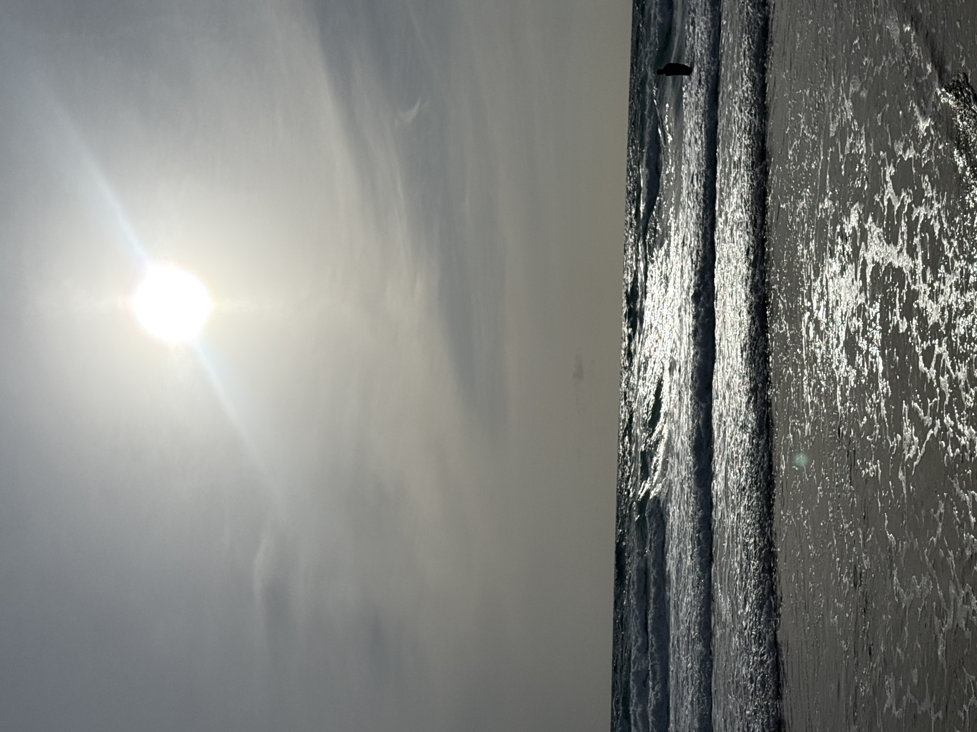 The image depicts a silhouetted figure standing in the ocean with waves breaking. The sun is high in the sky, casting a bright glare. The scene is likely Karon Beach, Karon, Thailand, known for its expansive shoreline and gentle waves. The water is a deep blue, and the sand is wet from the tide. The sky is a pale gray, with a few wispy clouds. The overall impression is one of tranquility and natural beauty.