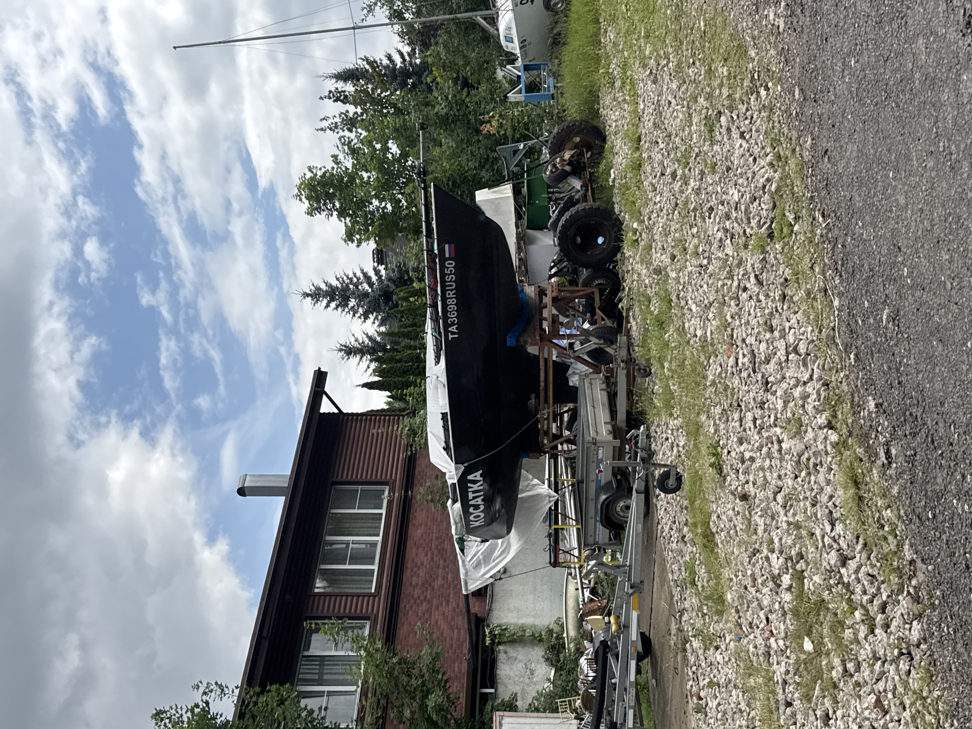 In Povedniki, Russia, a black sailboat, identified by the name "KOCATKA" and the registration "TA3698RUS50" with a Russian flag, rests on a trailer supported by a metal frame with large wheels. The boat is partially covered with a white tarp. In the background, a two-story wooden house with red siding and white-trimmed windows stands to the left, with a metal chimney on the roof. Several trees, including evergreens and deciduous trees, surround the area. To the right of the sailboat, another white boat is visible, and a tall mast indicates the presence of a sailboat in the distance. The ground is covered in a mixture of gravel and grass, with a paved surface visible in the foreground. The sky is partly cloudy with patches of blue. No people are present in the image.