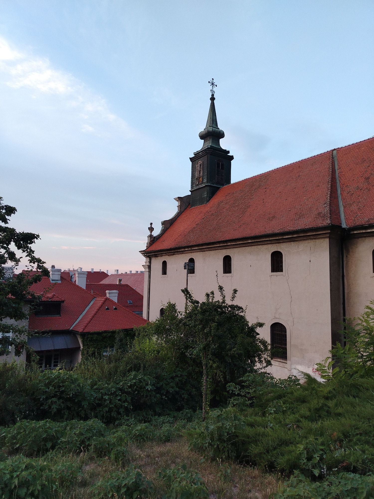This image shows a view of the Petrovaradin Fortress in Petrovaradin, Serbia. The focus is on a church with a distinctive steeple, characterized by its dark, elongated shape topped with a cross. The church building itself is light-colored with a red tiled roof and arched windows. Surrounding the church are other buildings with similar red roofs, indicating a dense urban or fortified area. Lush green trees and vegetation partially obscure the lower portions of the buildings and fill the foreground, suggesting a natural setting within or adjacent to the urban environment. The sky above is partly cloudy with hues of blue and pink, indicating it might be sunrise or sunset. No people are visible in the image.