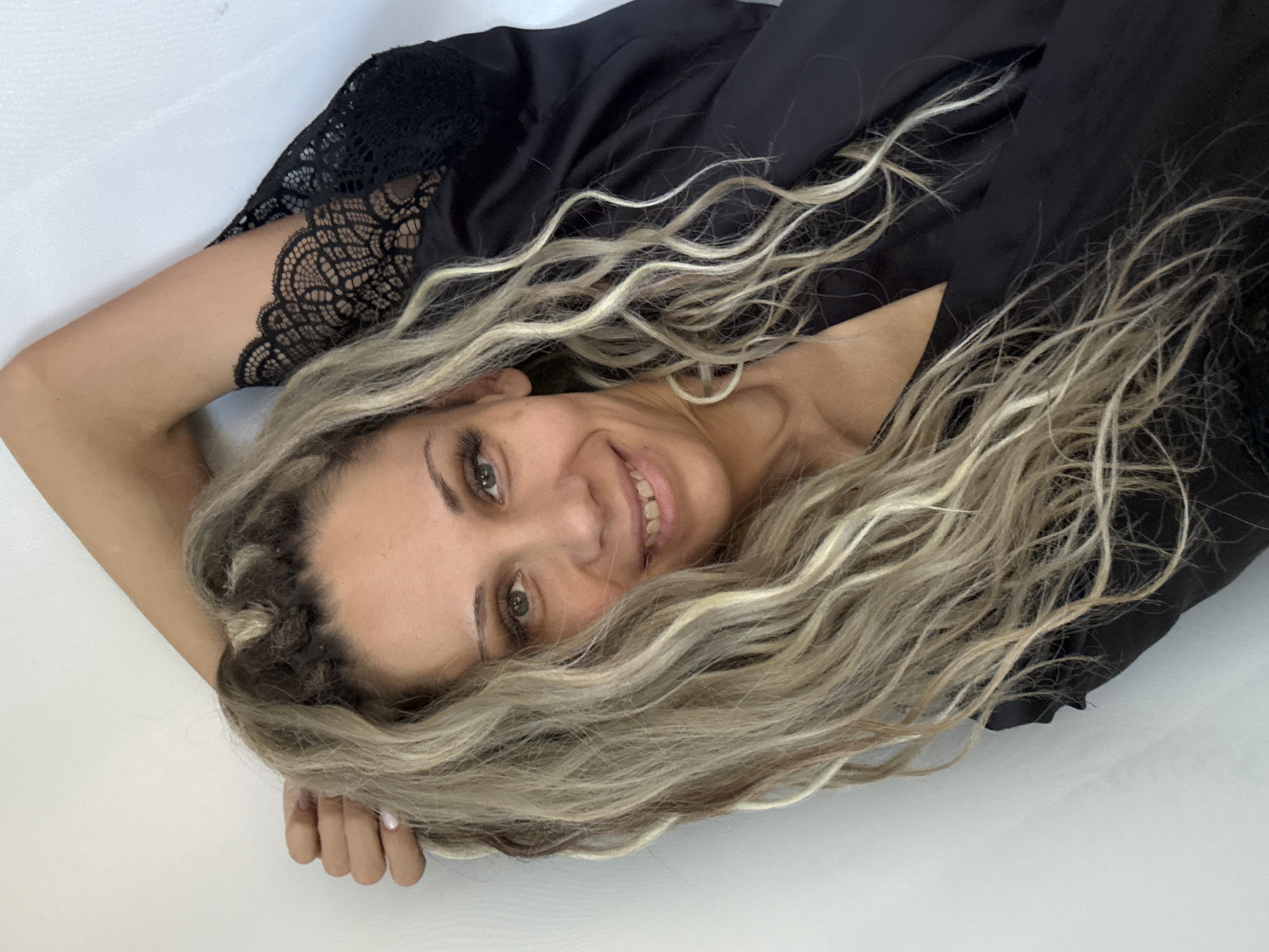 In Rusanovo, Russia, a woman with long, wavy blonde and brown hair is captured in a close-up, selfie-style portrait. She is wearing a black silk robe with lace trim and has her arm behind her head. The woman is looking directly at the camera with a smile, her eyes are green, and her teeth are slightly uneven. She has noticeable makeup, including long false eyelashes. The background is a plain white wall, providing a clean, uncluttered setting.