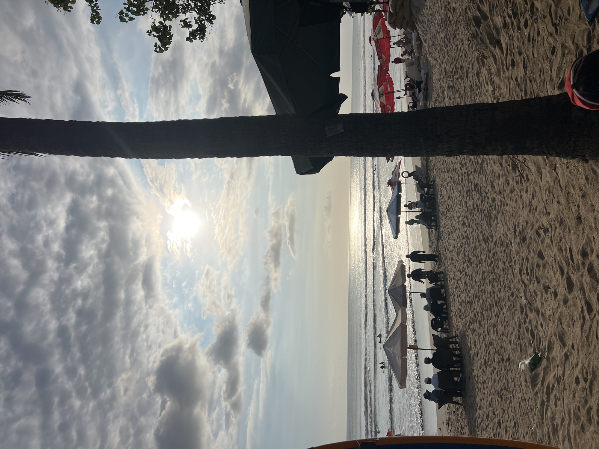 A beach scene in Kuta, Indonesia, features the sun low in the sky, casting a warm glow on the water and sand. The ocean has gentle waves, and several people are visible in the water, some with surfboards. On the sandy shore, beach umbrellas provide shade for lounge chairs and seating areas where small groups of people are gathered. A tall palm tree stands in the foreground, partially obscuring the view of a large umbrella structure. The sky is partly cloudy, with the sun shining through. The overall atmosphere is one of relaxation and leisure.