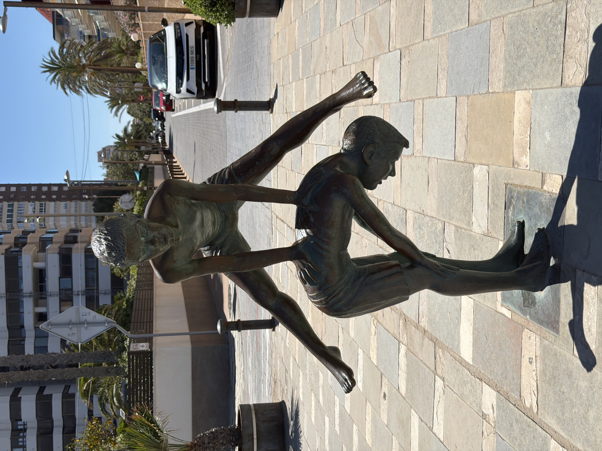 The image features a bronze sculpture of two boys stretching on a cobblestone pathway in Alicante, Spain. One boy is seated with legs extended, while the other stands facing him, both with arms outstretched. The sculpture is positioned on a square metal base, with visible shadow. A street scene in the background includes a multi-story building, palm trees, streetlights, and parked cars, including a white vehicle with the license plate "1089 NKS." A street sign and planters with vegetation are also visible. The sky is clear and blue.
