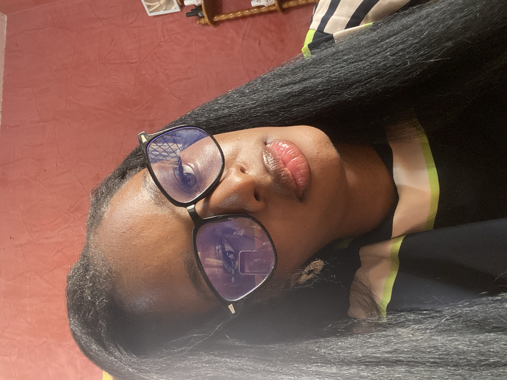 A close-up, selfie-style, waist-up shot of a young Black woman wearing black-rimmed glasses and a black, green, and white striped top is featured. She has long, straight black hair that is styled to frame her face and shoulders. The background is a textured, reddish-orange wall. The woman's face is slightly turned towards the camera, and her lips are glossy and slightly parted. The glasses reflect a pattern that resembles a cross-hatch design, and also a smaller reflection of what appears to be a smartphone screen. To the right of the woman, a wooden shelf unit with several bottles, possibly nail polish, is visible against the wall. The lighting in the image is somewhat diffused, creating highlights on her skin and glasses. The overall impression is of a casual portrait.