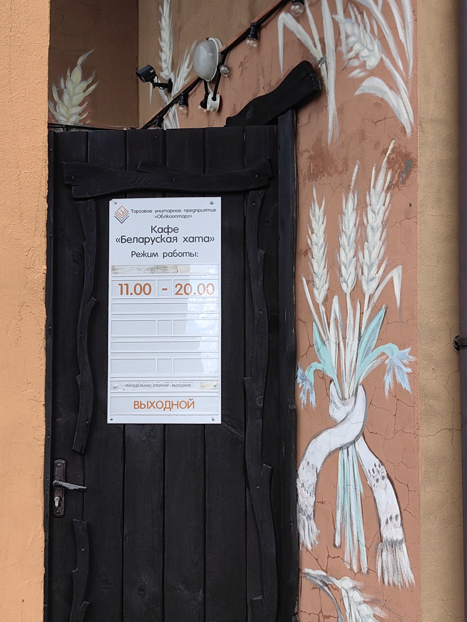 The image shows a dark wooden door with a white sign indicating the operating hours of a cafe. The sign reads "Торговое унитарное предприятие «Облконпторг»", "Кафе «Беларуская хата»", and "Режим работы: 11.00 - 20.00". Below the hours, it says "ПОНЕДЕЛЬНИК, ВТОРНИК - ВЫХОДНОЙ" and "ВЫХОДНОЙ". The door is on the left side of the image, and a painted mural of wheat stalks decorates the wall to the right. The cafe is located in Myadzyel, Belarus.