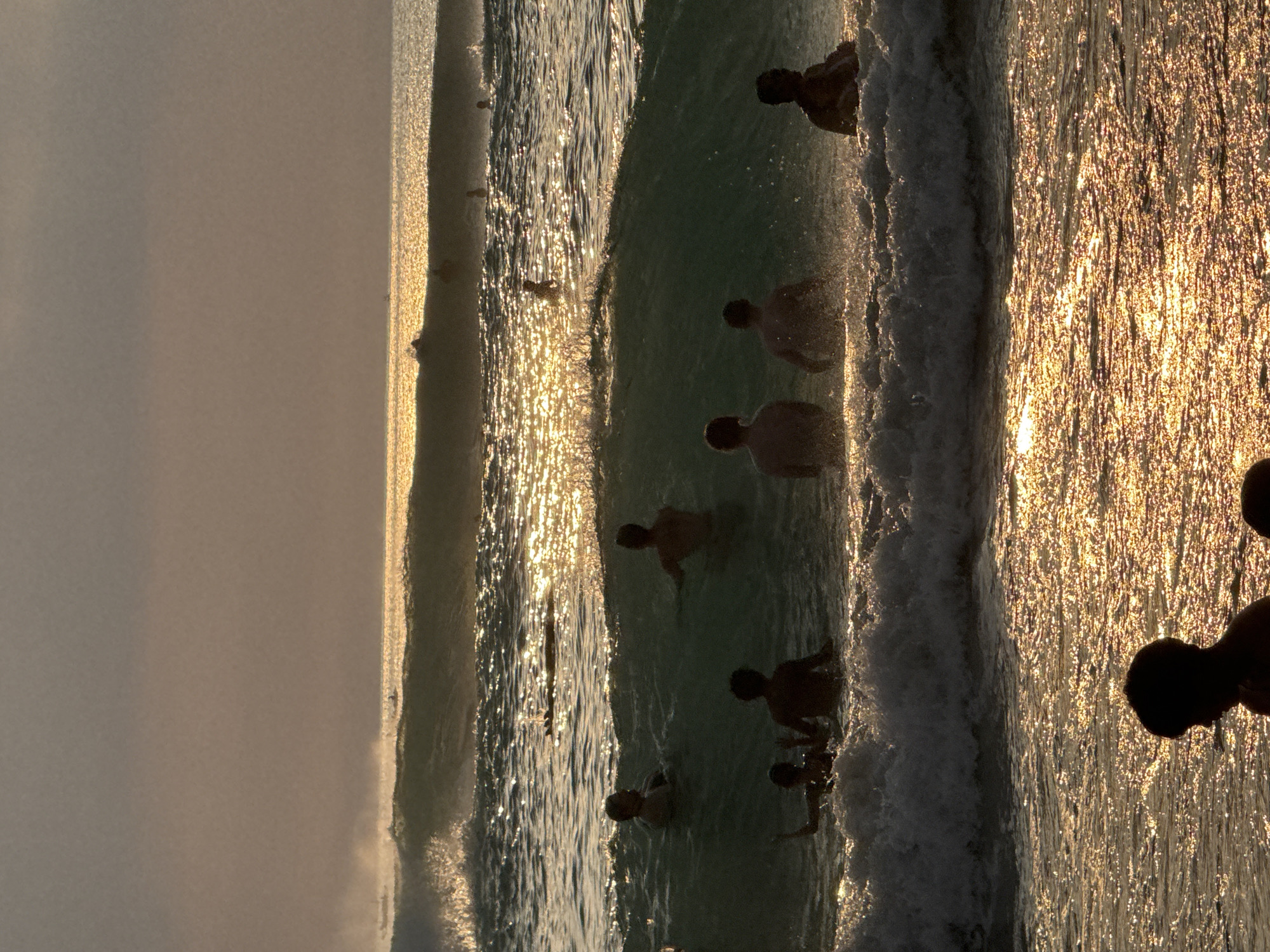 The image depicts a silhouetted group of people swimming and playing in the ocean at Kuta Selatan, Indonesia, during sunset. The sun's golden light reflects off the water, creating a shimmering effect. Several individuals are visible in the water, some standing, others partially submerged. Waves are breaking, with foam and spray visible. In the background, more people can be seen further out in the water, and the horizon is marked by the meeting of the sea and a hazy sky. The overall atmosphere is one of leisure and recreation in a coastal environment.