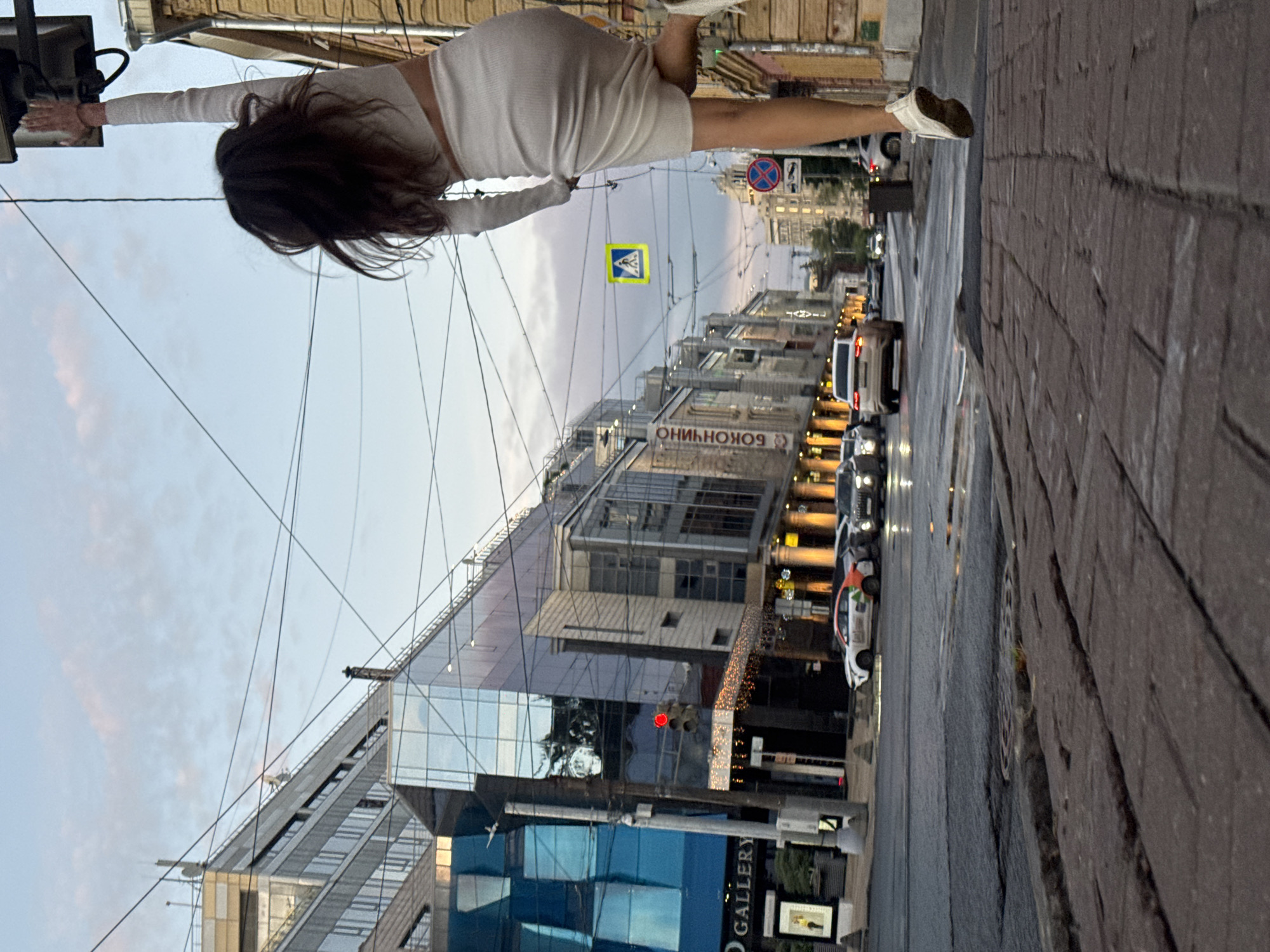 The image captures a dynamic street scene in Nizhny Novgorod, Russia. In the foreground, a woman in a white knit top and skirt is suspended mid-air, reaching up towards a light fixture. Her body is angled, with her right leg extended and her left leg bent. Her dark hair is blurred, indicating movement. She is dressed in casual attire, with white sneakers.
The background depicts a city street with buildings on either side. The left side features a modern building with a glass facade, reflecting the sky and surrounding structures. A sign reading "GALLERY" is visible on this building. A traffic light with a red signal is positioned near the entrance of this building. Further down the street, another building displays signage that appears to be in Cyrillic, possibly "БОКОНЧИНО". The right side of the street has an older building with yellow brickwork.
The street is wet, suggesting recent rain, and reflects the streetlights and traffic lights. Several cars are on the road, including a white car with colorful decals and a dark SUV. A pedestrian crossing sign is visible in the middle ground, a yellow square with a blue pictogram of a person walking. The sky is overcast with hints of pink and orange, indicating either dawn or dusk. Overhead wires crisscross the sky, common in urban environments. The pavement in the foreground is made of red brick pavers.
