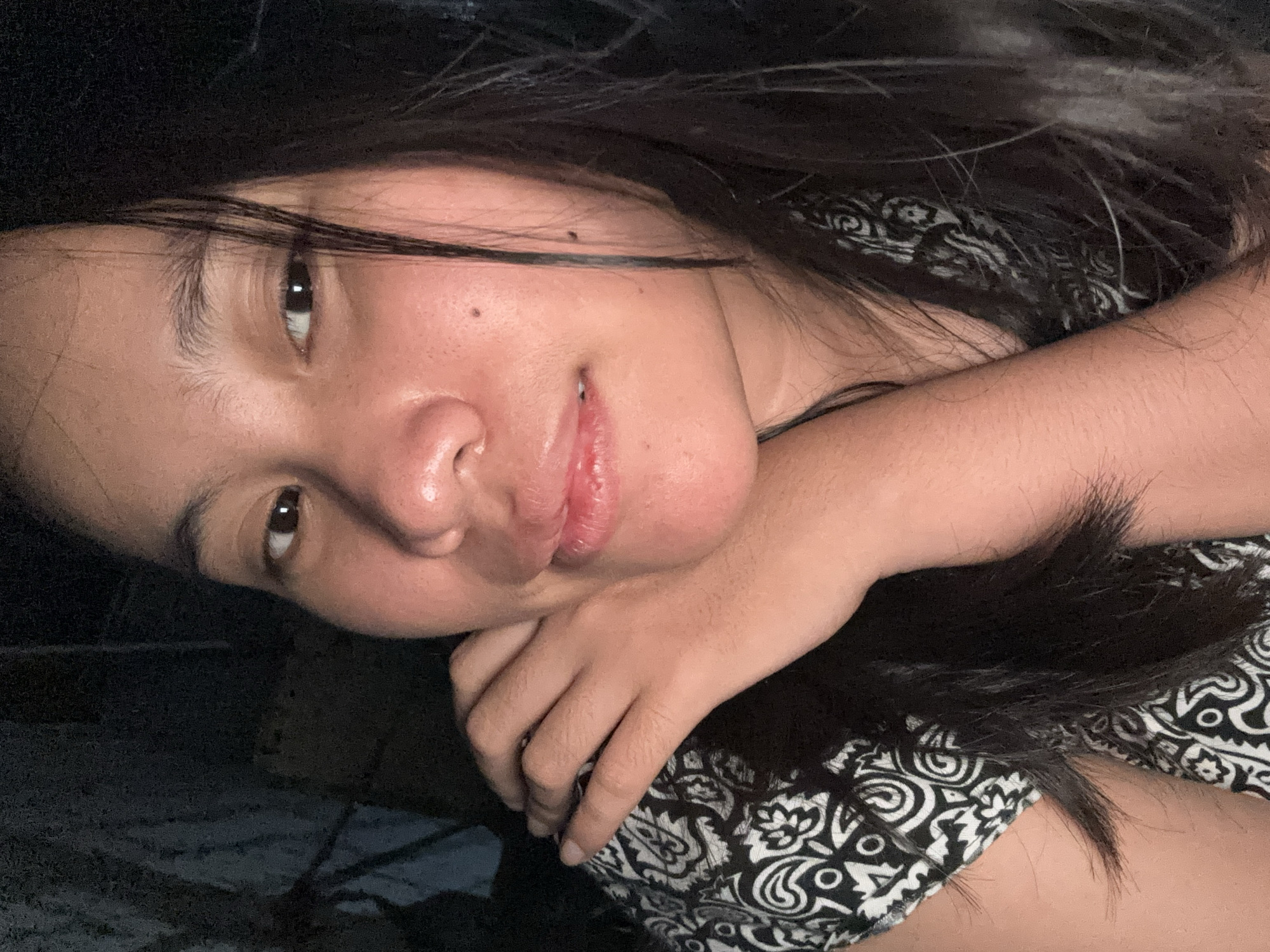 A close-up, head-and-shoulders, selfie-style photograph shows a young woman from East Jakarta, Indonesia, resting her chin on her left hand. Her right arm is bent, and her hand is positioned near her face, resting on her left shoulder. She is wearing a black and white patterned shirt. Her dark hair is partially obscuring her face and shoulders. The background is dark and indistinct, suggesting an indoor setting with low lighting. The woman's gaze is directed towards the camera.