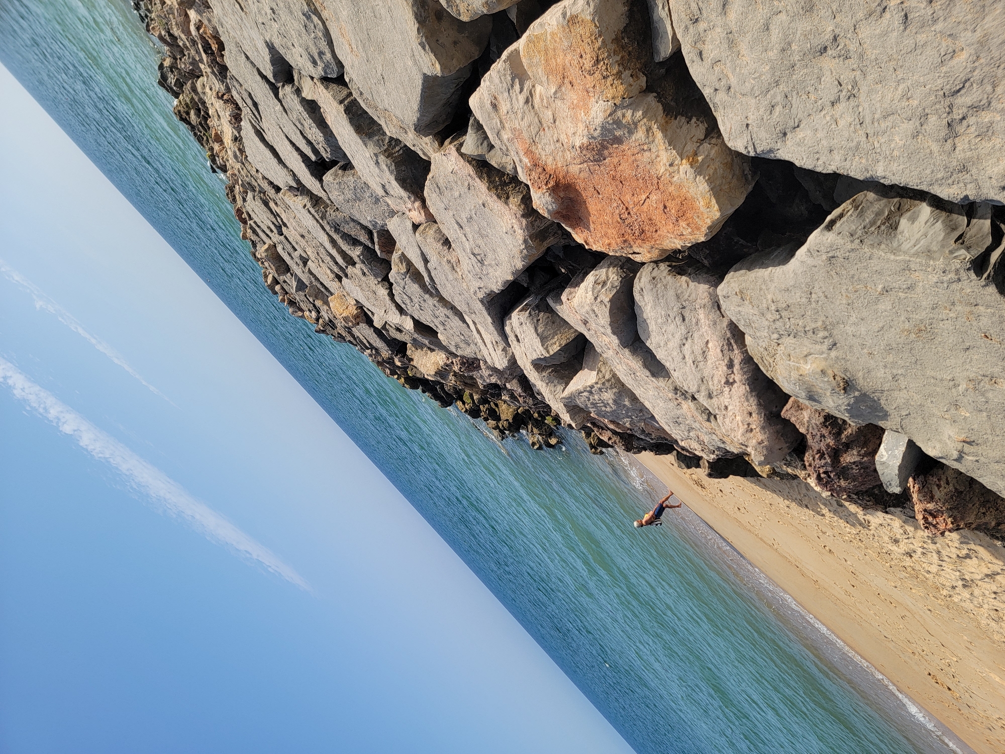 This media file depicts a person standing on the sandy shore of a beach, adjacent to a rocky breakwater. The ocean is calm with turquoise waters, and the sky is a clear, pale blue with a wispy white cloud formation stretching across it. The sun appears to be high in the sky, casting shadows from the rocks, suggesting it is midday. The scene is one of tranquility, with the vast expanse of the sea and sky emphasizing a sense of openness.
The individual, a male, is wearing dark shorts and a light-colored t-shirt or tank top. He is facing away from the viewer and towards the water, with his arms slightly raised and his body in a pose that could indicate contemplation or anticipation of entering the water. The sand is light brown and appears dry further from the water's edge, with gentle waves lapping at the shore. The breakwater is constructed of large, irregularly shaped gray rocks, some showing hints of reddish-brown mineral deposits. The rocks are piled high, creating a stark, natural barrier against the sea.
The image is captured from a slightly elevated, diagonal perspective, with the horizon line running from the upper left to the lower right of the frame. This orientation adds a dynamic feel to the otherwise still scene. There is no visible text in the media file. The overall impression is a serene moment at the seaside, captured during a bright, sunny day.