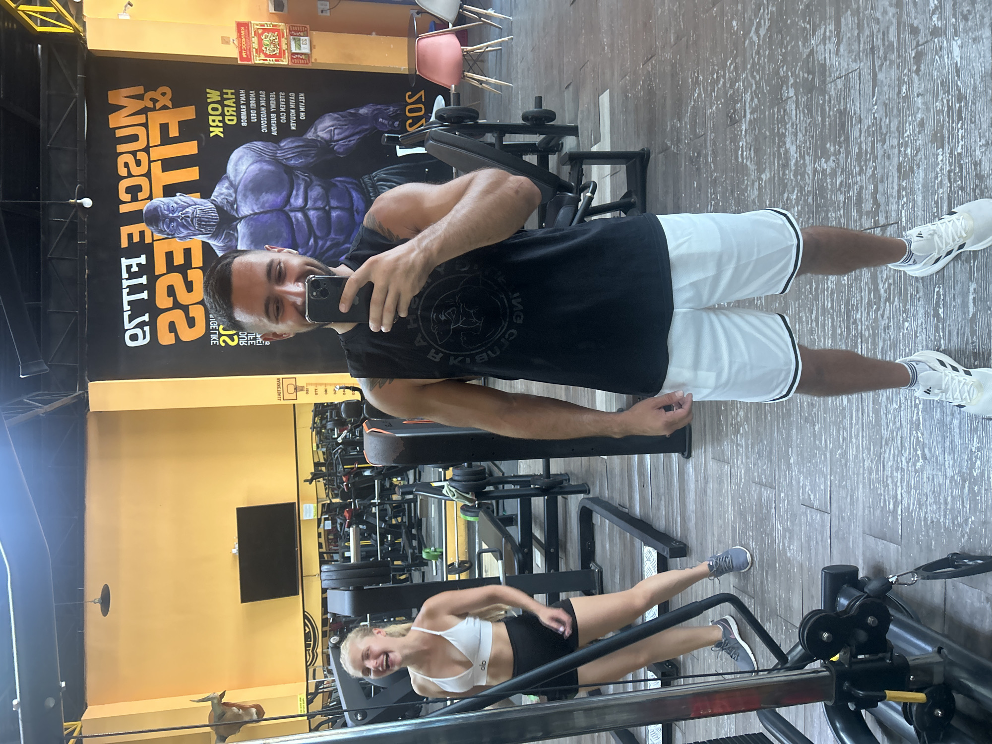 The image captures a selfie taken by a man in a gym, likely located in Nha Trang Ward, Vietnam, given the presence of Vietnamese text on a poster. The man, dressed in a black tank top and white athletic shorts, is holding a smartphone to take the photo. He has short dark hair and a beard, and he is smiling.
Behind him, a woman in a white sports bra and black athletic shorts is visible, laughing. She appears to be working out. The gym environment is evident with various weightlifting equipment, including barbells, weight plates, and a weight machine.
A large mural featuring a muscular, purple-skinned character, possibly Thanos from the Marvel comics, adorns the wall behind the man. The mural has text in English and what appears to be Vietnamese. The gym has yellow walls and a wooden floor with visible wear. Other elements in the background include gym equipment, chairs, and a wall-mounted television.