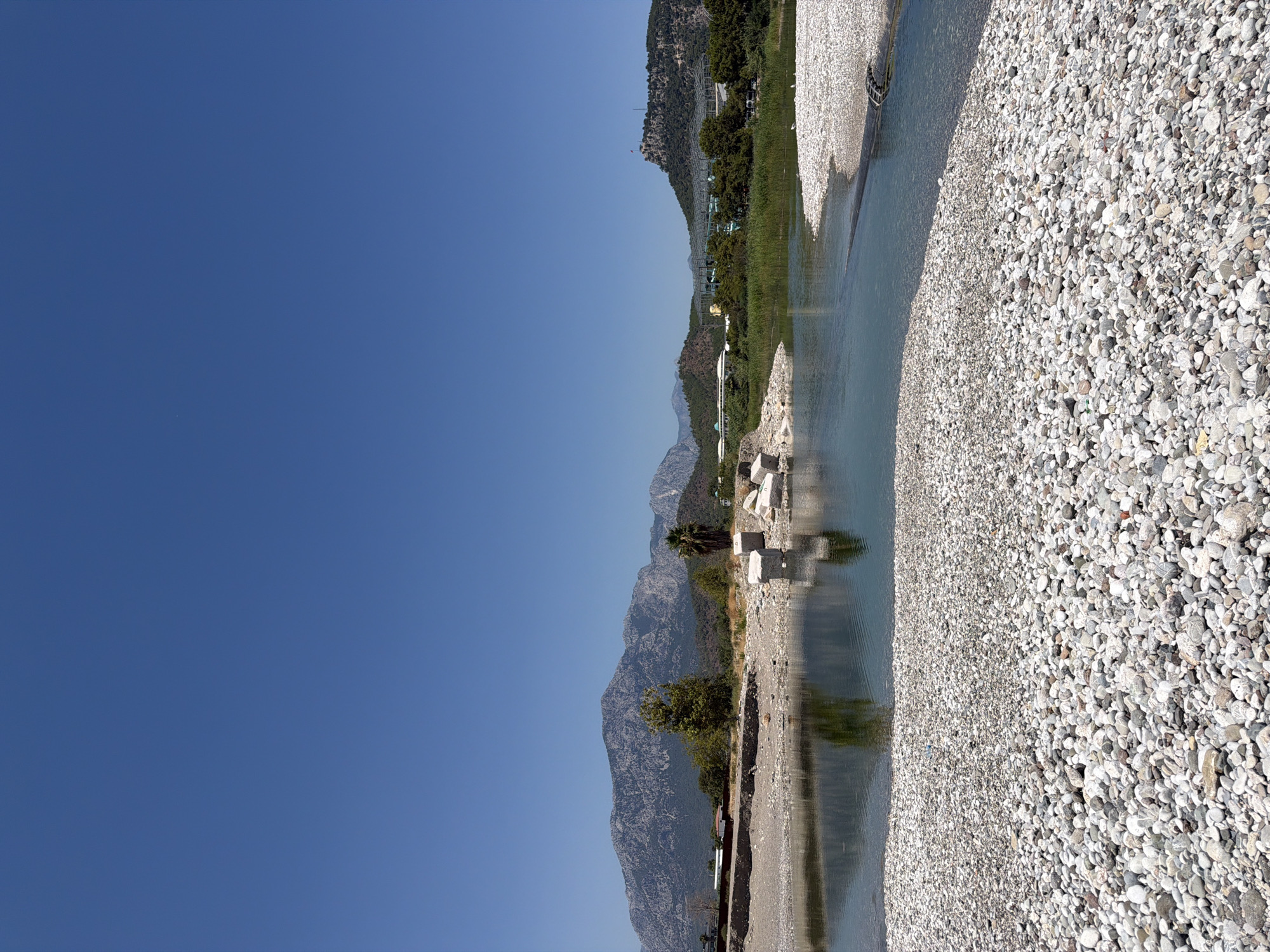 The image depicts a wide, clear blue sky over a natural landscape. In the mid-ground, a body of water, likely a river or a lake, reflects the surrounding environment. The banks of the water are lined with smooth, light-colored pebbles. Beyond the water, a thin line of green vegetation runs horizontally, separating the water from a rocky, mountainous terrain that dominates the background. The mountains appear rugged and sparsely vegetated. To the right of the frame, a building complex with a distinctive architectural style, possibly a resort or entertainment venue, is visible nestled among the trees. A flag flies atop a small hill to the far right. Scattered white concrete blocks are visible along the pebbled shore in the mid-ground, suggesting some form of construction or remnants of an older structure. The overall scene conveys a tranquil, sun-drenched day in a coastal or mountainous region.