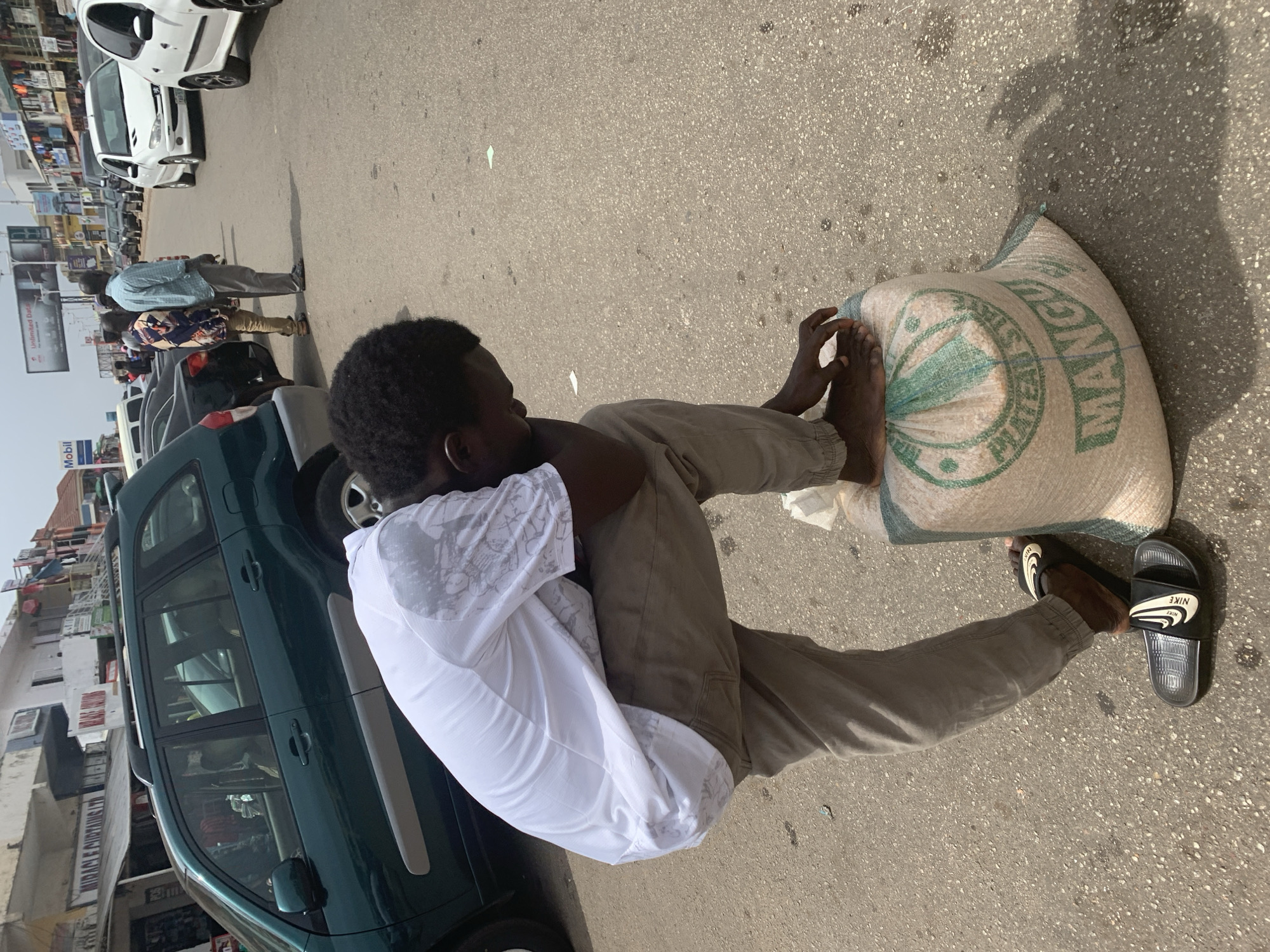 A man is sitting on a bag on the street in Jos, Nigeria. He is Black, wearing a white shirt and tan pants. His feet are bare, resting on the bag. Next to the bag, he is wearing Nike slides. The bag is light brown and has a green circular design on it with the word "WANGU" printed on it. The street is asphalt. Behind the man is a green car and other cars parked along the street. Buildings are visible on the left side of the image.