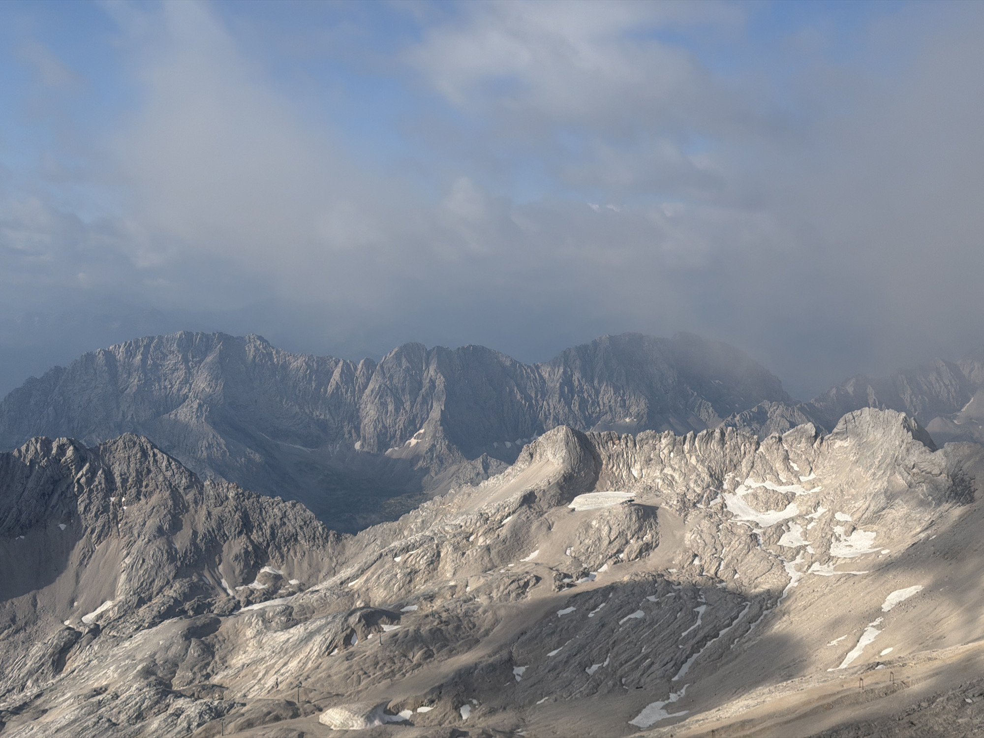 The image is a high-angle, wide shot of a rugged mountain range under a cloudy sky. The mountains are predominantly rocky and gray, with patches of snow visible on some slopes. The foreground shows a steep, rocky terrain with dramatic shadows cast by the uneven surface. In the middle ground, more mountain peaks rise, some with jagged edges and others appearing more rounded. The background reveals a chain of distant, hazy mountains shrouded in mist or clouds. The sky is a mix of light blue and gray clouds, with sunlight breaking through in some areas, creating illuminated sections on the mountains. There are no people or man-made structures clearly visible, except for what might be distant ski lift poles in the lower right quadrant. The scene appears to be a natural, mountainous landscape, likely in a cold or alpine environment. While not explicitly identifiable from the image alone, the prompt specifies the location as Grainau, Germany, which is known for its proximity to the Zugspitze, Germany's highest peak, and the surrounding Alps.