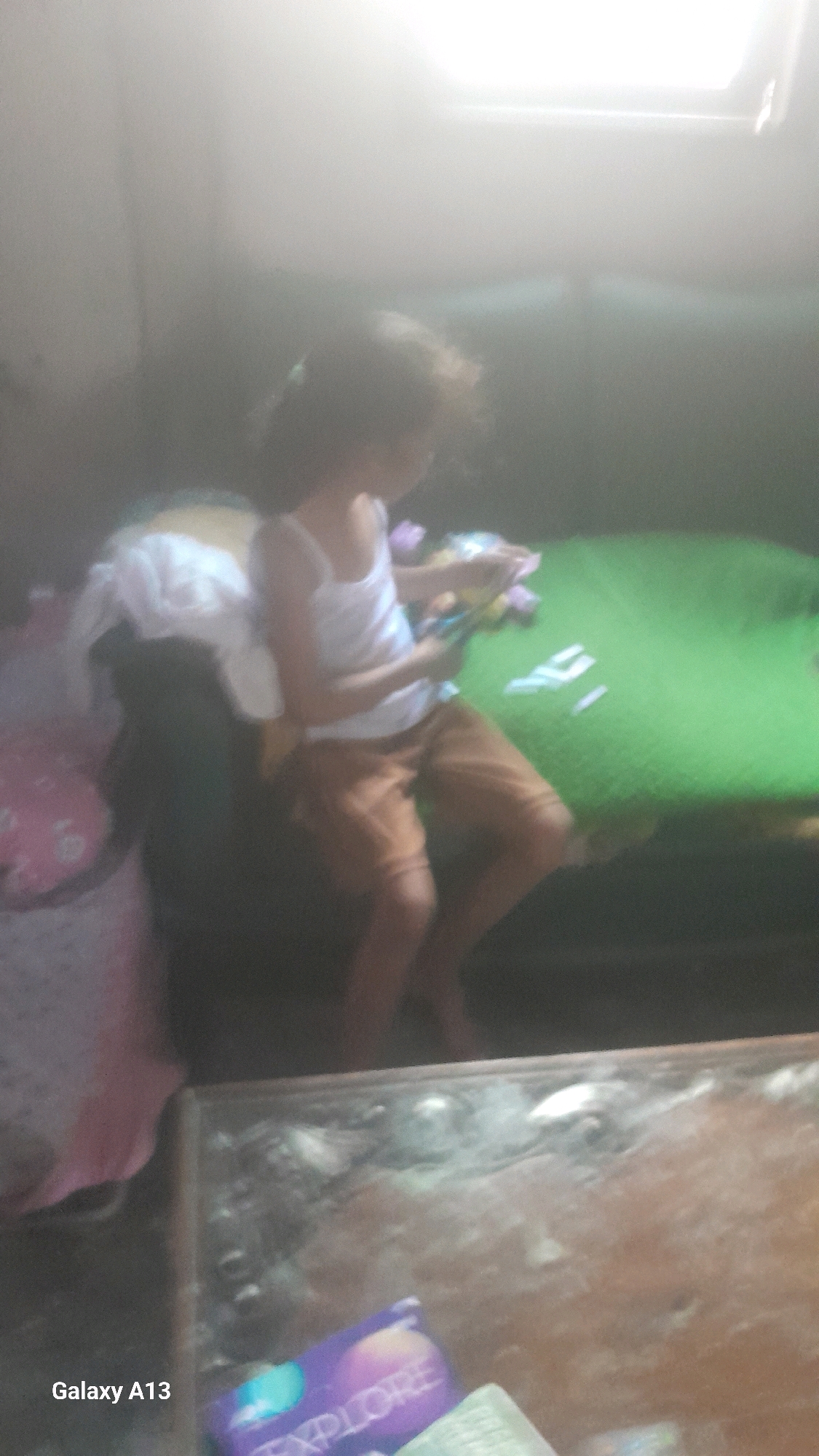 In the city of Ngelo, Indonesia, a child sits on a dark object, possibly a chair or cushion, in what appears to be a dimly lit room. The child is wearing a white tank top and brown shorts and is holding what looks like art supplies, possibly crayons or markers, and a piece of paper. In front of the child is a green surface, possibly a mat or bed, with several white rectangular objects arranged on it. To the left of the child, a pink object with white floral patterns is visible, and behind the child, a white fabric is draped over the dark object. The foreground of the image shows a table with some debris or dust on its surface, and a purple object with the word "EXPLORE" printed on it, along with the text "Galaxy A13". The overall lighting suggests an indoor environment, possibly a home, with a bright light source emanating from above, possibly a skylight or overhead lamp.