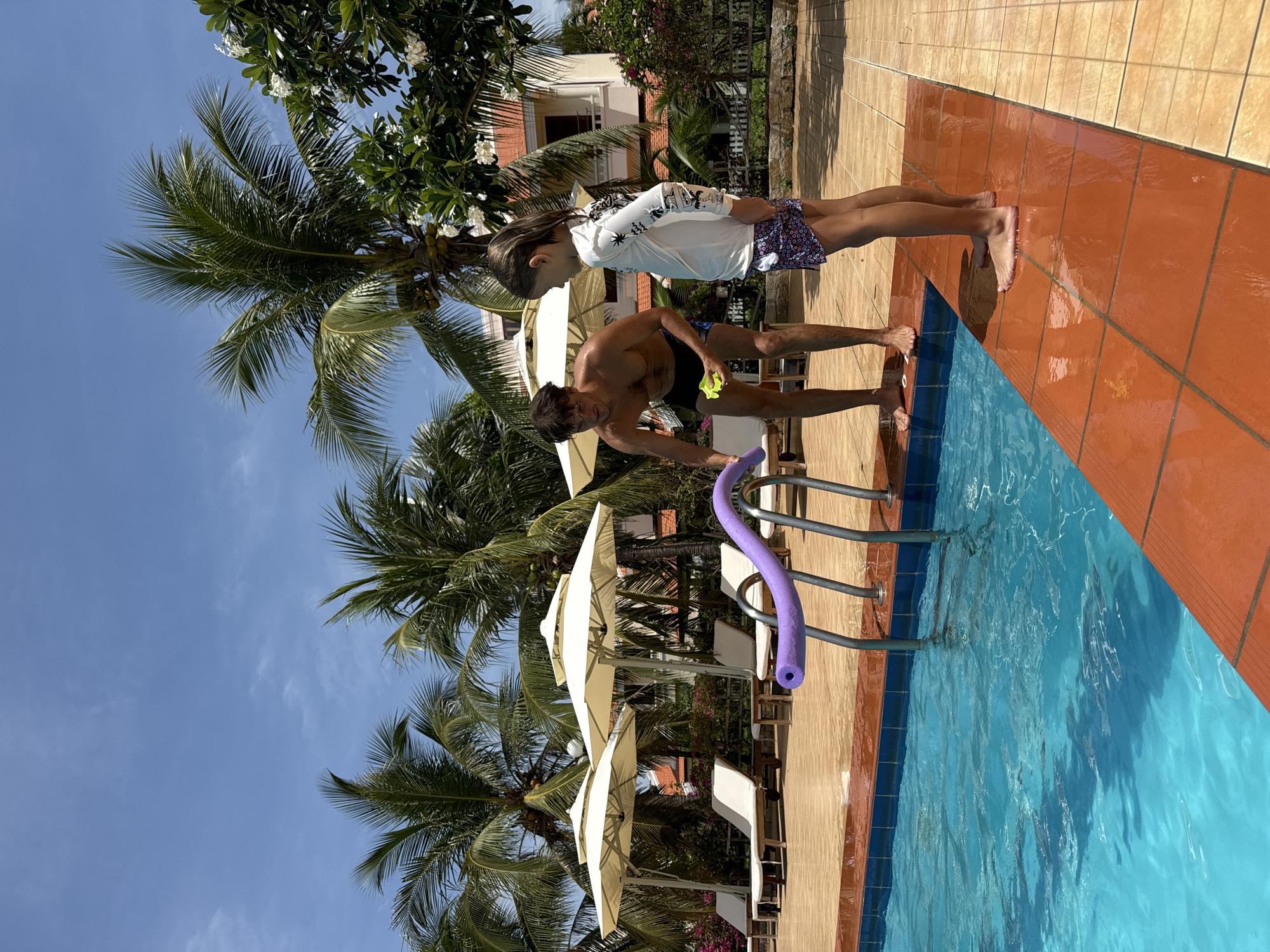 Scene in Phu Thuy Ward, Vietnam, near a pool. A man in dark swimwear leans towards a pool ladder, holding a purple pool noodle and a yellow object. A child in a white long-sleeved shirt and patterned shorts stands beside him. The pool is surrounded by orange and beige tiling. Palm trees and white patio umbrellas are visible in the background. The sky is blue with scattered clouds.