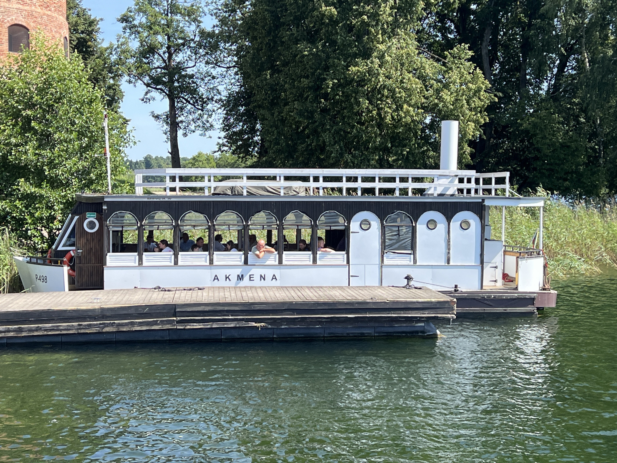 The image shows a white boat named "AKMENA" docked at a wooden pier on a body of water, likely a lake in Trakai, Lithuania. The boat has a dark-colored upper deck with arched windows and a white lower hull. Several passengers are visible inside the boat, seated and looking out. The background features lush green trees, a tall brick tower to the left, and a blue sky. The water is green and slightly rippled. The pier is made of dark wooden planks.