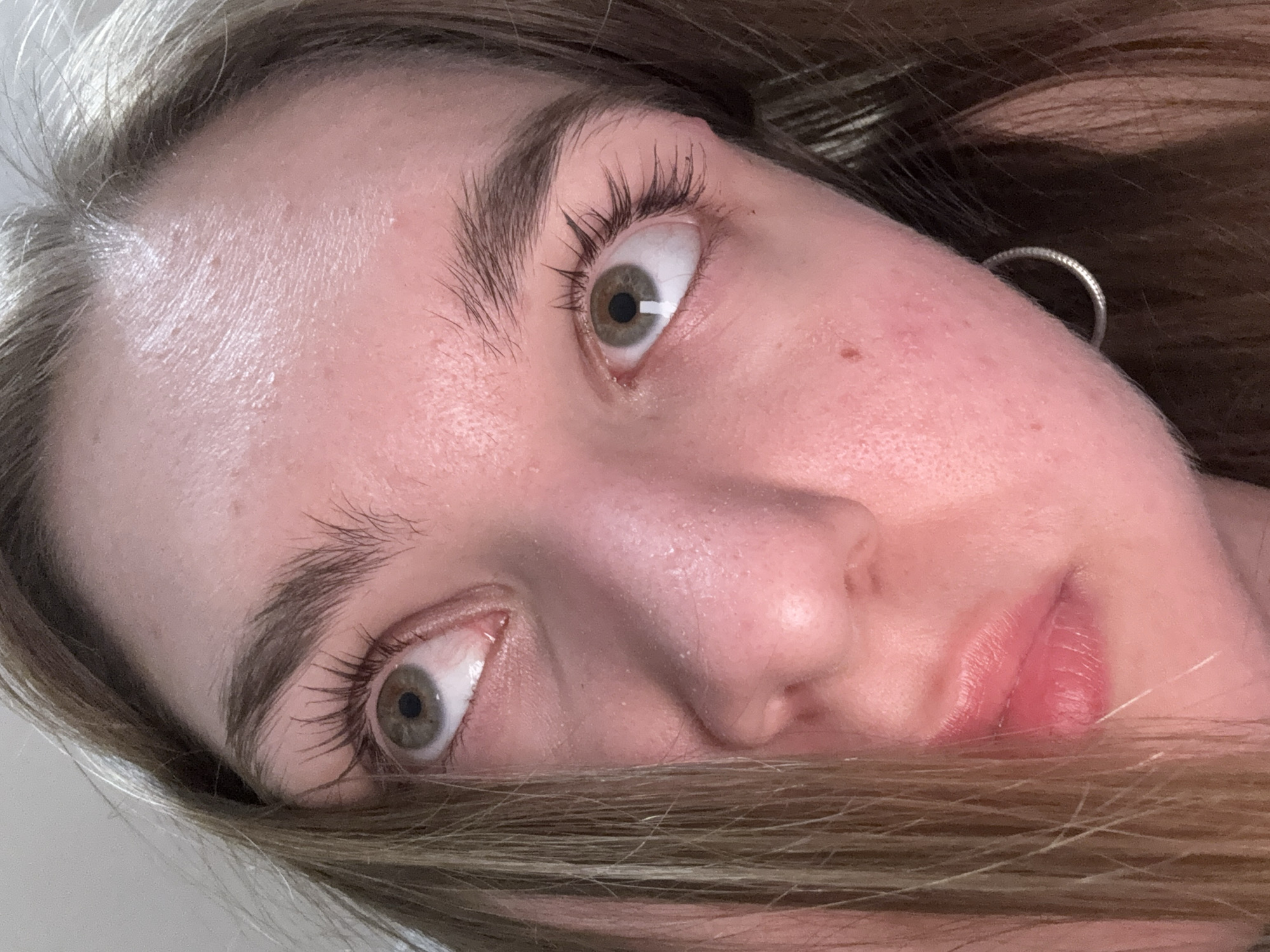 A close-up, angled portrait of a fair-skinned person with light brown hair. The person's face is tilted, with one eye appearing larger than the other due to perspective. The person has light-colored eyes and their eyelashes are clearly visible. The skin appears smooth with some visible pores and a few minor blemishes. Part of an earring is visible on the right side of the image. Some strands of hair are obscuring the lower part of the face. There is no evidence to suggest the location is Perm, Russia. The environment is indistinct, with a plain background partially visible on the left side.