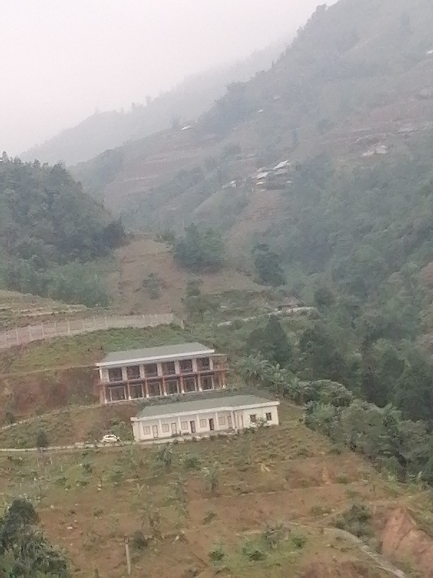 The image shows a mountainous landscape with two buildings situated on a hillside. The buildings are of different sizes and designs. The larger building appears to be a two-story structure with multiple windows, while the smaller building is a single-story structure. A white car is parked on the slope between the two buildings. The surrounding environment includes trees, vegetation, and terraced fields. The overall setting suggests a rural or mountainous region. The sky is overcast, and the atmosphere appears misty or foggy.