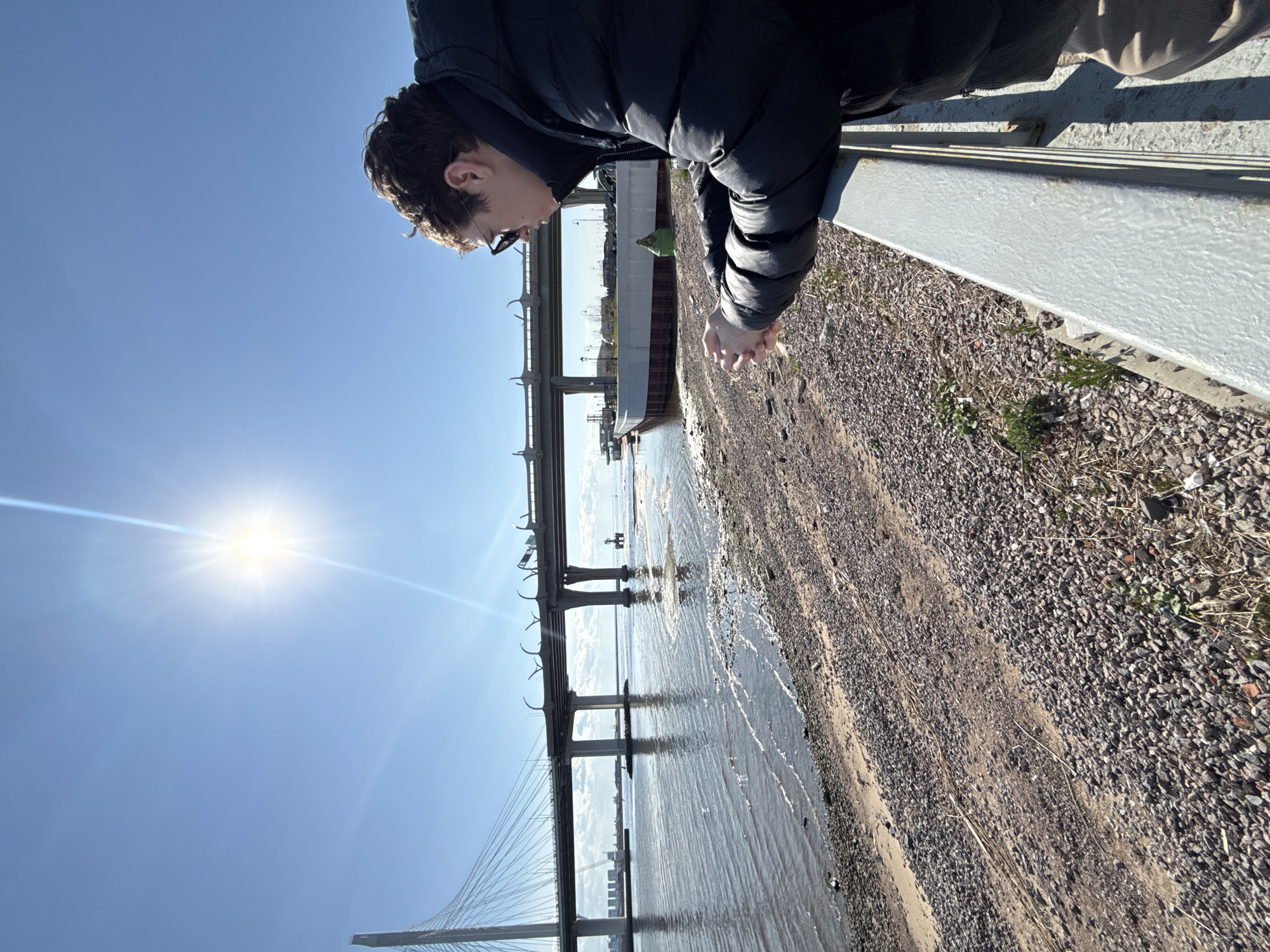 A person is leaning over a concrete railing, looking down at a sandy shoreline. Beyond the shoreline is water, with a large ship partially visible in the distance. Bridges, including a cable-stayed bridge, span the water. The sky is bright blue with a shining sun. The scene appears to be near the Gulf of Finland in Saint Petersburg, Russia, based on the bridge structure and coastline. The immediate foreground consists of gravel and sparse vegetation. The person is wearing a dark jacket.