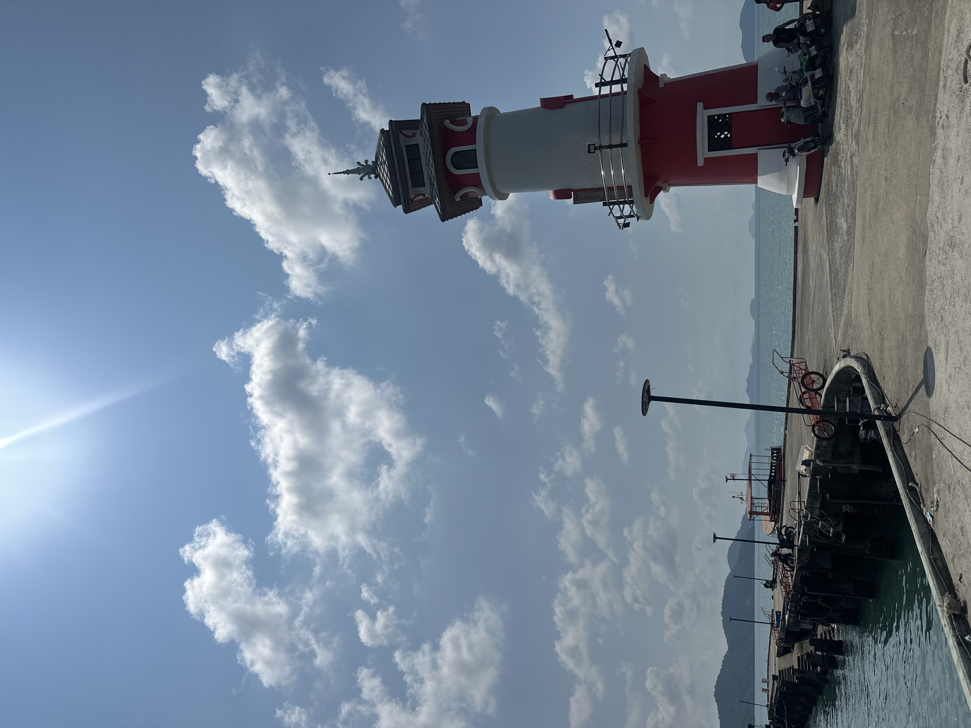 A low-angle, medium shot taken in Baan Bai Lan, Thailand. The image features a red and white lighthouse, a concrete pier, a body of water, and a sky with clouds. The lighthouse stands on the pier, and several parked motorbikes are near the lighthouse's base. The water stretches to the horizon, with mountains visible in the distance. Several black light posts are on the pier.