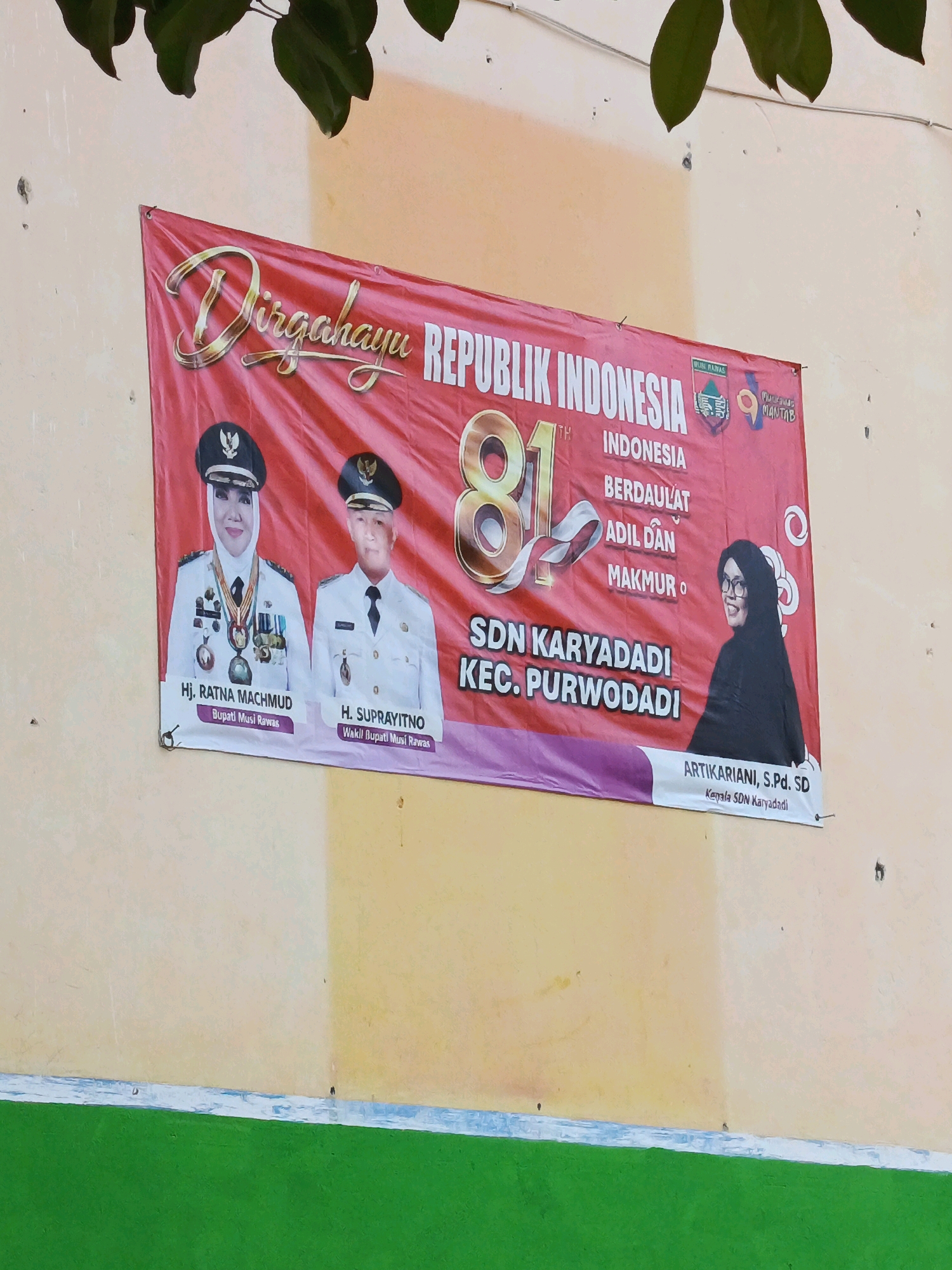 A red banner is displayed on a cream-colored wall. The banner reads "Dirgahayu REPUBLIK INDONESIA 81st INDONESIA BERDAULAT ADIL DAN MAKMUR O". There are portraits of three individuals on the banner. On the left is Hj. Ratna Machmud, Bupati Musi Rawas, wearing a white uniform and a hijab. Next to her is H. Supriyatno, Wakil Bupati Musi Rawas, also in a white uniform. On the right is a woman identified as ARTINAKARIANI, S.Pd. SD, Kepala SDN Karyadadi, wearing a black hijab and glasses. The banner also features logos and the text "SDN KARYADADI KEC. PURWODADI". The wall is painted in alternating shades of cream and light yellow, with a bright green band at the bottom. Foliage from trees hangs over the top of the banner.
