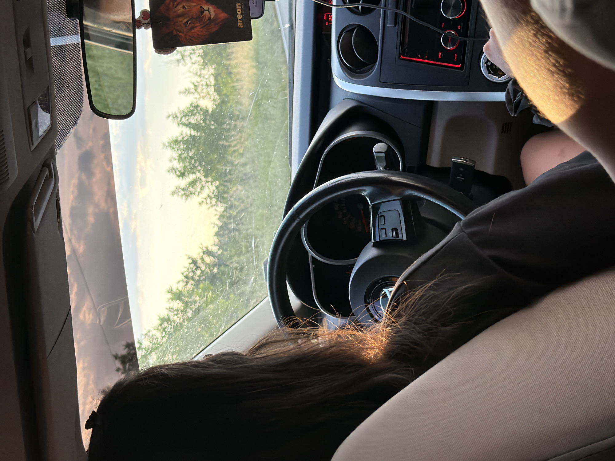 The interior of a car is depicted. A person with long, dark hair is sitting in the passenger seat. The driver's hands are visible, and they appear to be interacting with the car's dashboard. A rear-view mirror hangs above. The car is moving along a road that passes Byal Izvor, Bulgaria, as can be seen through the windshield. The scene is bathed in natural light.