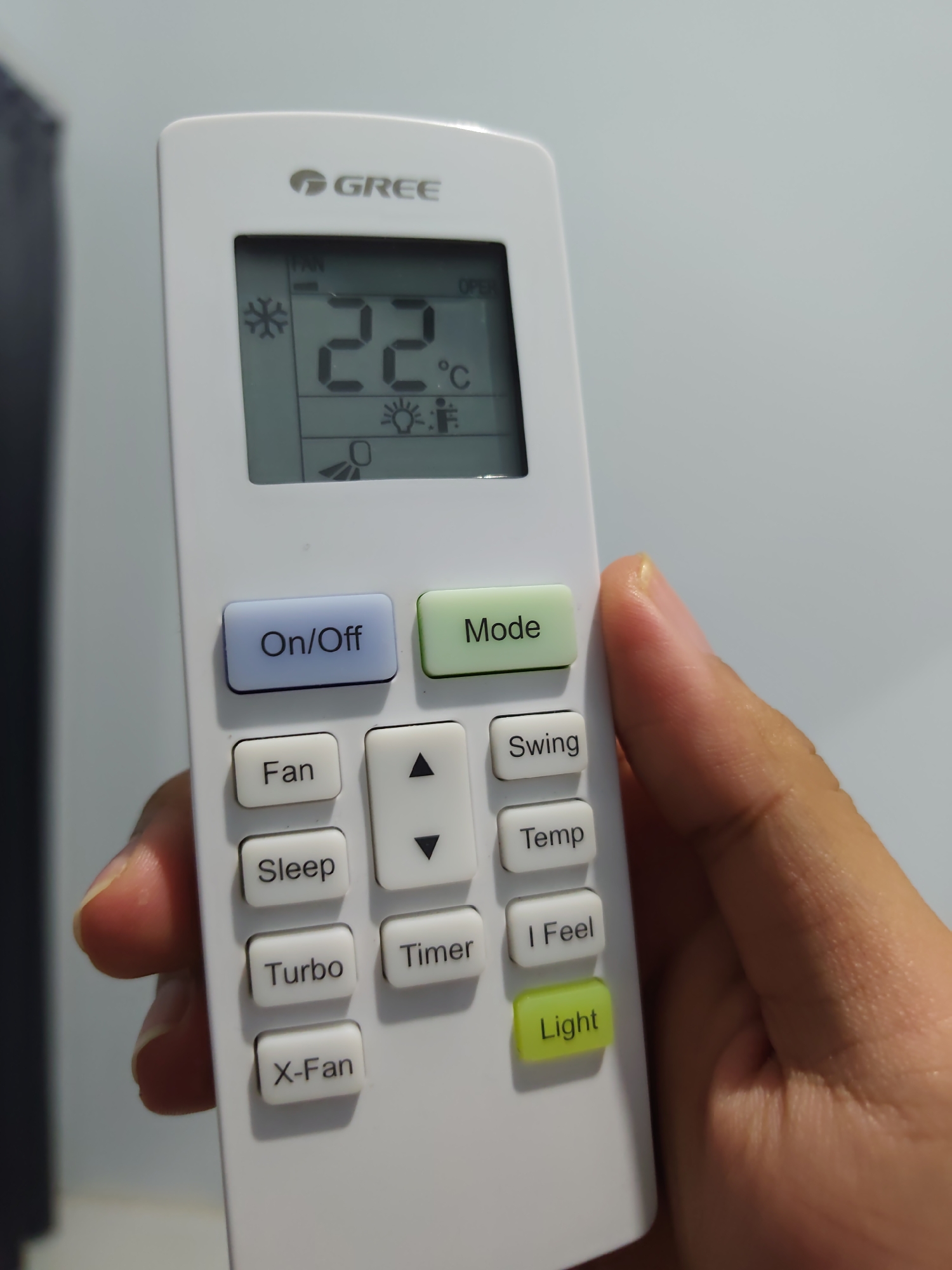 A hand holds a white Gree brand air conditioner remote control against a light gray wall. The remote has a digital display showing "22°C" and various icons indicating settings such as a snowflake for cooling, a light bulb, and a fan. Below the display are buttons labeled "On/Off", "Mode", "Fan", "Sleep", "Turbo", "X-Fan", "Swing", "Temp", "Timer", "I Feel", and "Light". The hand is positioned to press a button on the remote. The image is taken in Pamotan, Indonesia.