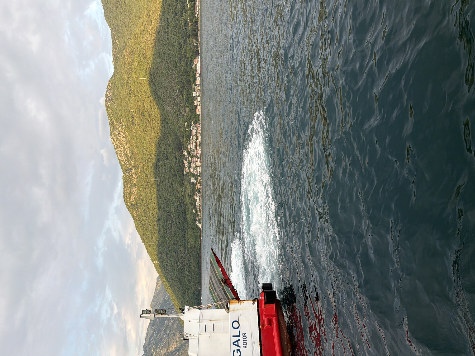 A ferry boat, identified by the markings "GALO KOTOR," is seen from its stern as it moves through the water, leaving a wake of white foam. The ferry has a red hull and a white superstructure. A ramp, partially raised, is visible at the stern. The boat is on a body of water, likely a bay or strait, with a lush green, mountainous coastline in the background. A town with white and red-roofed buildings is nestled along the shore at the base of the mountains. The sky above is partly cloudy, with soft sunlight illuminating the scene. The location is Đurići, Montenegro. There are no people visible on the ferry or in the immediate vicinity.