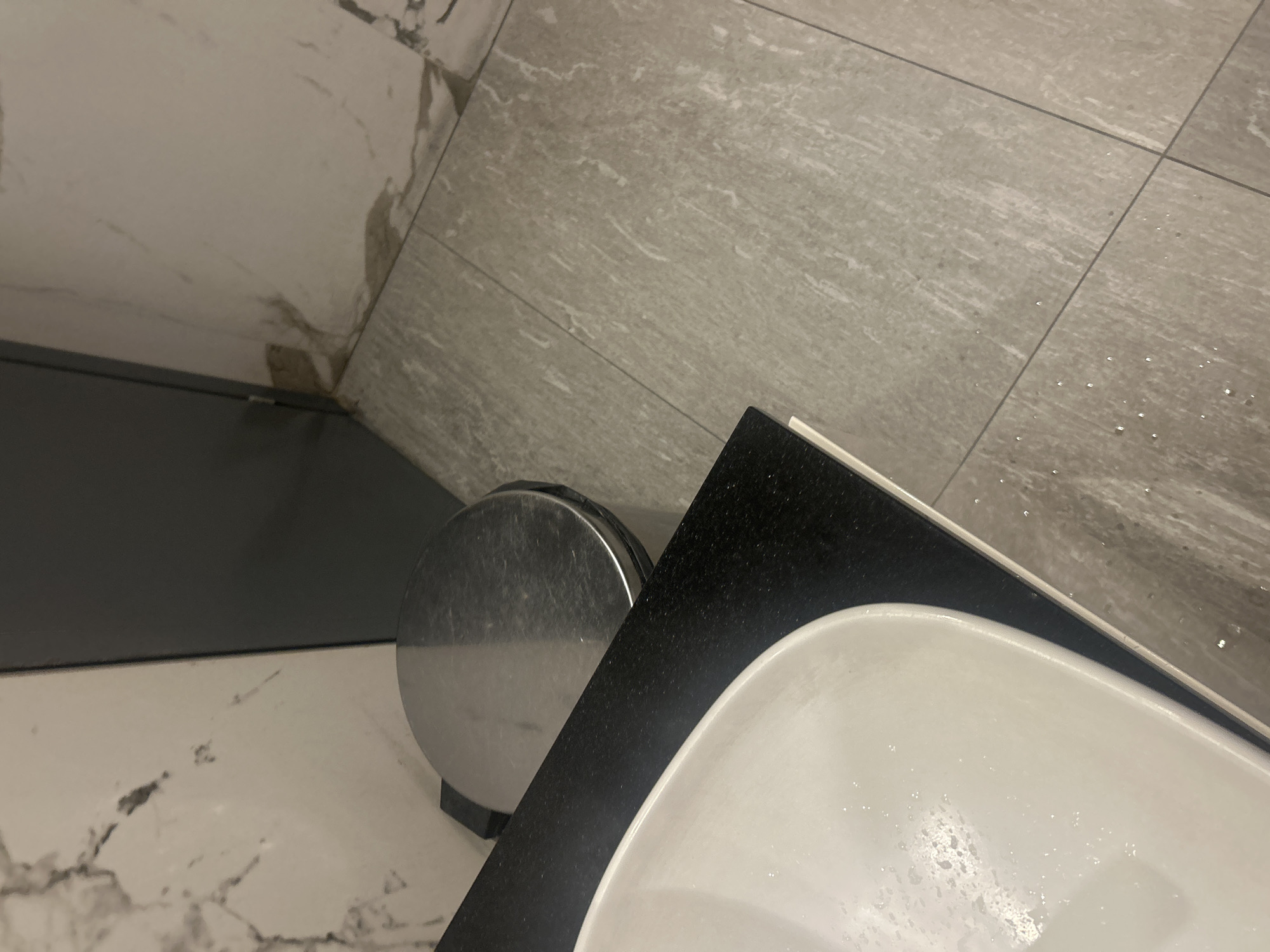 A close-up, high-angle shot shows a modern bathroom sink with a white basin and a black countertop. To the left of the sink is a round, metallic trash can. The floor is tiled with a gray, wood-grain pattern, and there are small droplets of water visible on the tiles. The wall behind the sink features large, light-colored marble-patterned tiles. A dark gray panel runs along the bottom of the wall. The scene suggests a clean and contemporary bathroom environment, possibly in Plovdiv, Bulgaria. No people or text are visible, and the lighting indicates an indoor setting, likely during daytime.