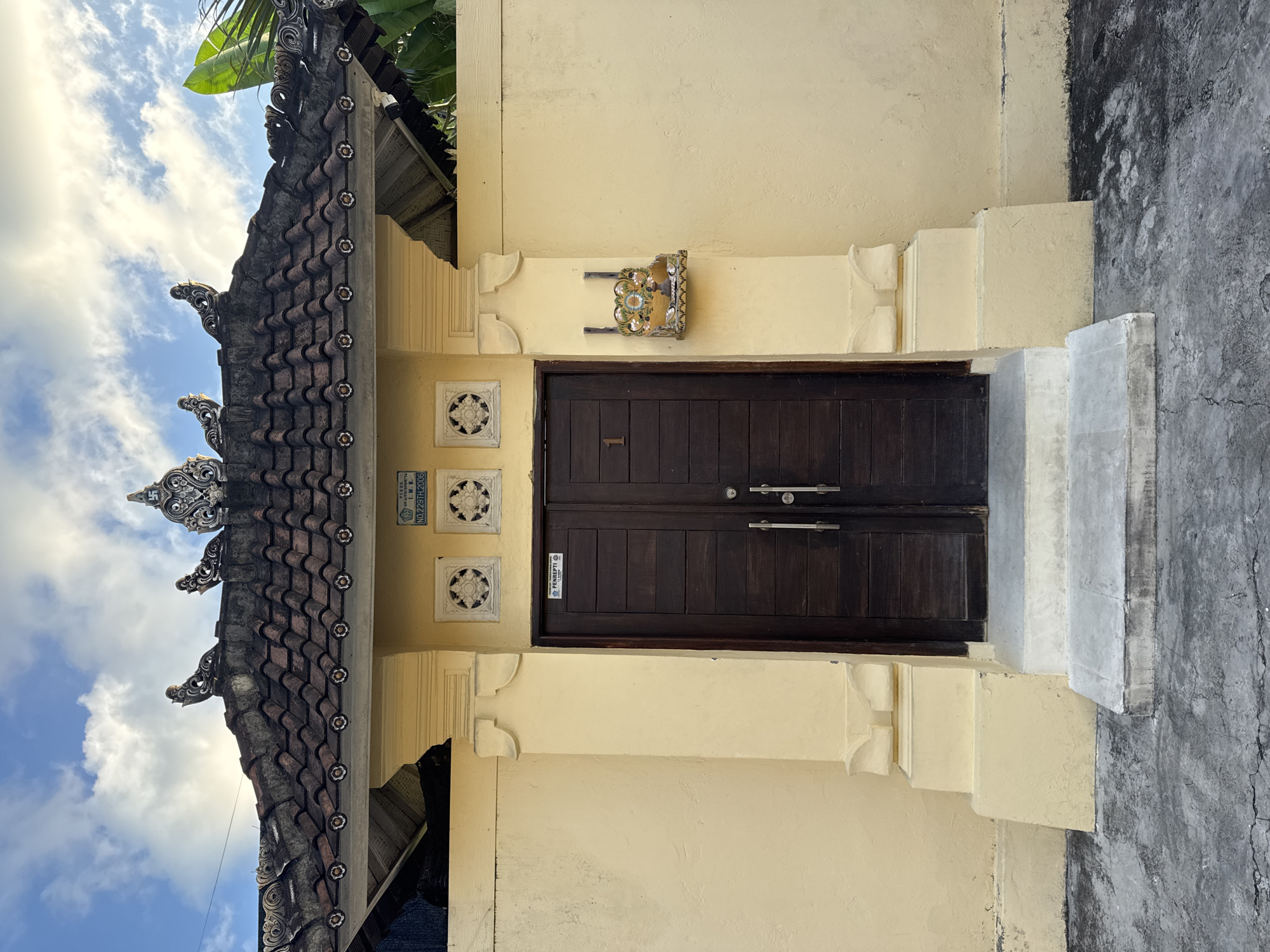 A detailed view of a traditional Balinese door in Kuta, Indonesia. The dark brown, double wooden door is positioned centrally, featuring silver handles and a small notice. Above the door is a small, ornate shelf. Decorative, floral-patterned tiles are set into the yellow wall on the left side of the door. The roof section on the left side displays intricate, dark brown tiling and elaborate carvings, reaching towards a blue sky with scattered clouds and a green palm frond. A set of concrete stairs and a concrete wall are on the right of the door.