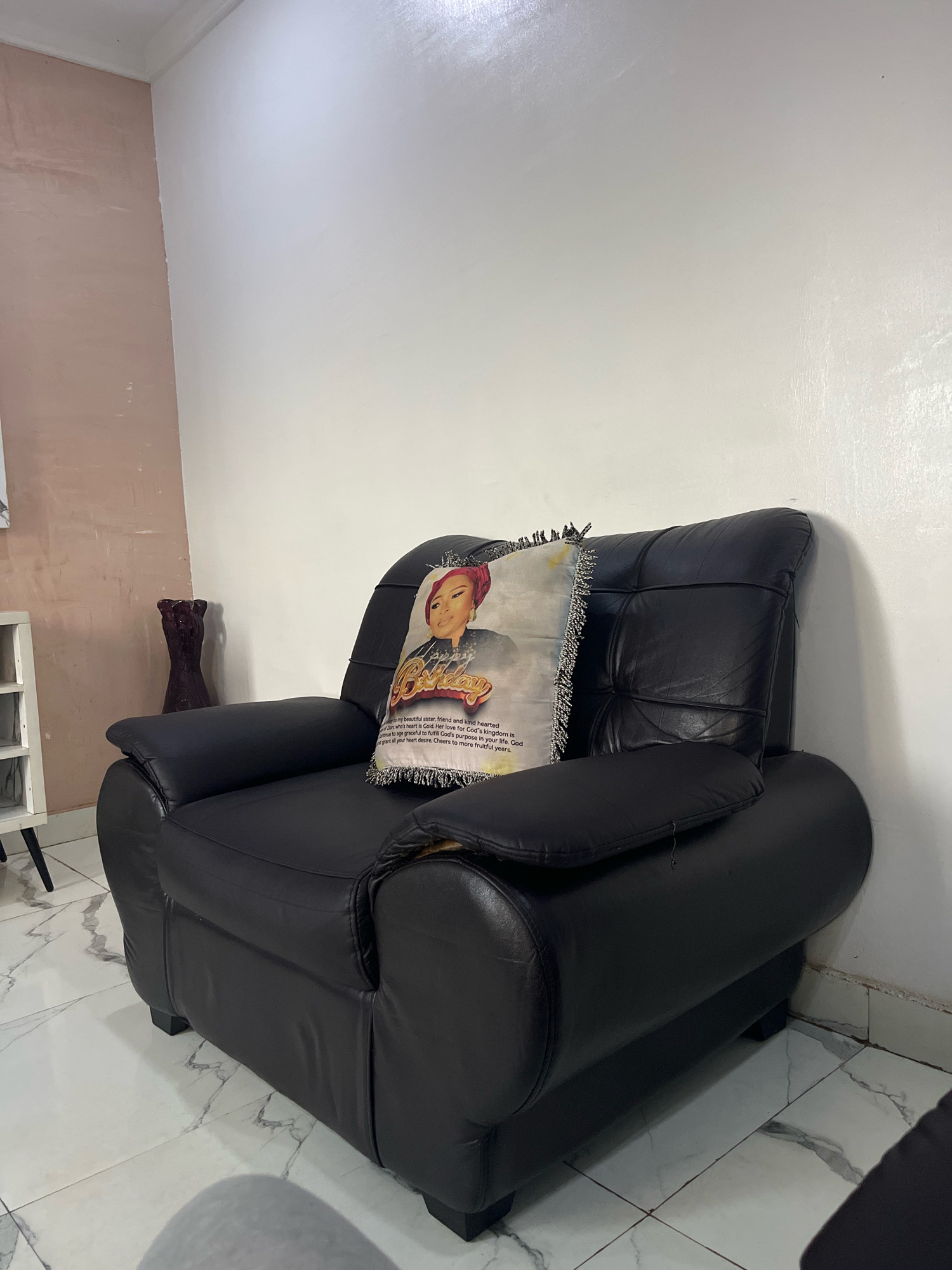 The image shows a black leather armchair with a decorative pillow placed on it. The pillow features a portrait of a woman with a red headscarf and the word "Birthday" in large font, along with text below it that appears to be a religious message. The armchair is situated in a room with white walls and tiled floors. To the left of the armchair, a portion of a white shelf unit is visible, and behind it, a dark, vase-like object stands against a wall painted in a dusty rose color. The overall setting suggests an indoor environment, possibly a living room. The image does not contain any specific visual cues to pinpoint its location within Jos, Nigeria.