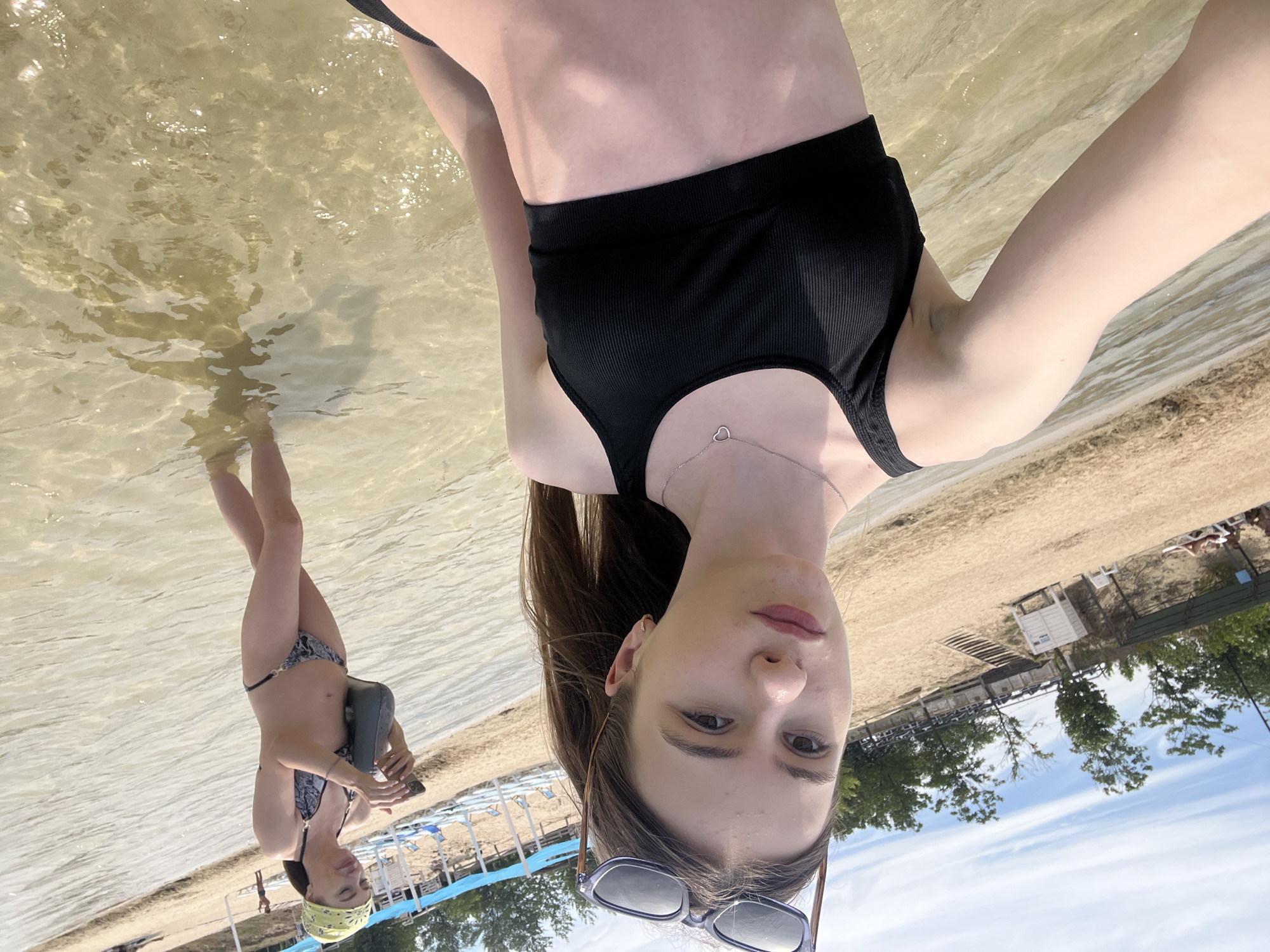 This image was taken at the Yelyzavetivka Reservoir, located in the city of Yelyzavetivka, Ukraine. The scene is a beach area with shallow, clear water. In the foreground, a young woman is taking a selfie, wearing a black bikini top and sunglasses perched on her head. She is standing in the water, with the sandy beach and trees visible in the background. Behind her, another woman in a patterned bikini and a yellow bandana is walking in the water, looking at her phone. Beach chairs and umbrellas are set up on the shore. The sky is partly cloudy.