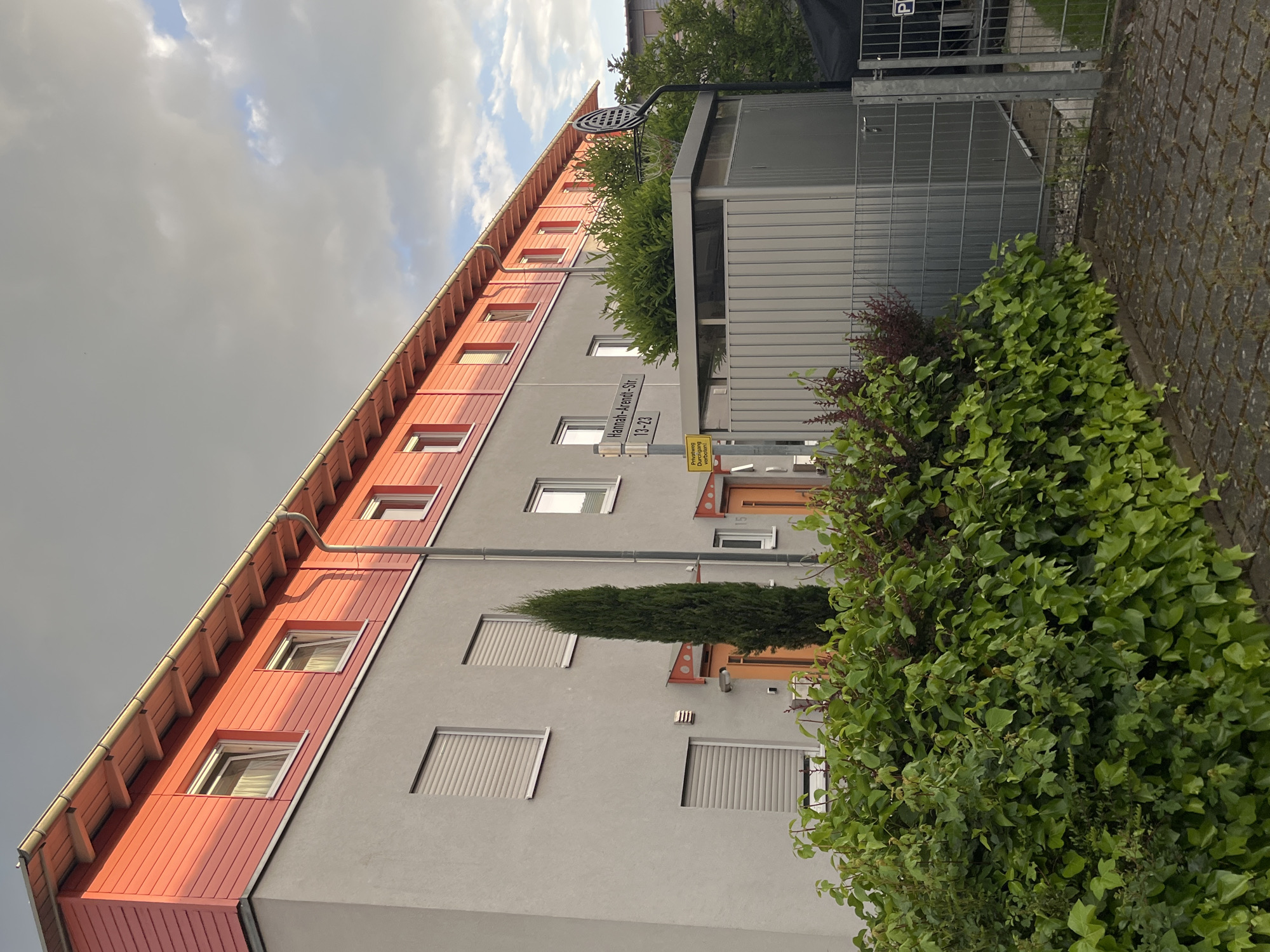 The image depicts a low-angle view of a multi-story building with a reddish-orange facade and a light grey stucco exterior. The building features several windows with white frames, some of which have partially closed blinds. A street sign indicating "Hannah-Arendt-Str. 13-23" is visible, confirming the location in Landau in der Pfalz, Germany. The sky is overcast, with a mix of light and dark clouds. Green foliage and bushes are prominently featured in the foreground, obscuring part of the building's entrance. A metal structure or enclosure stands next to the building. The ground is paved with grey stones. There are no people visible in the image.