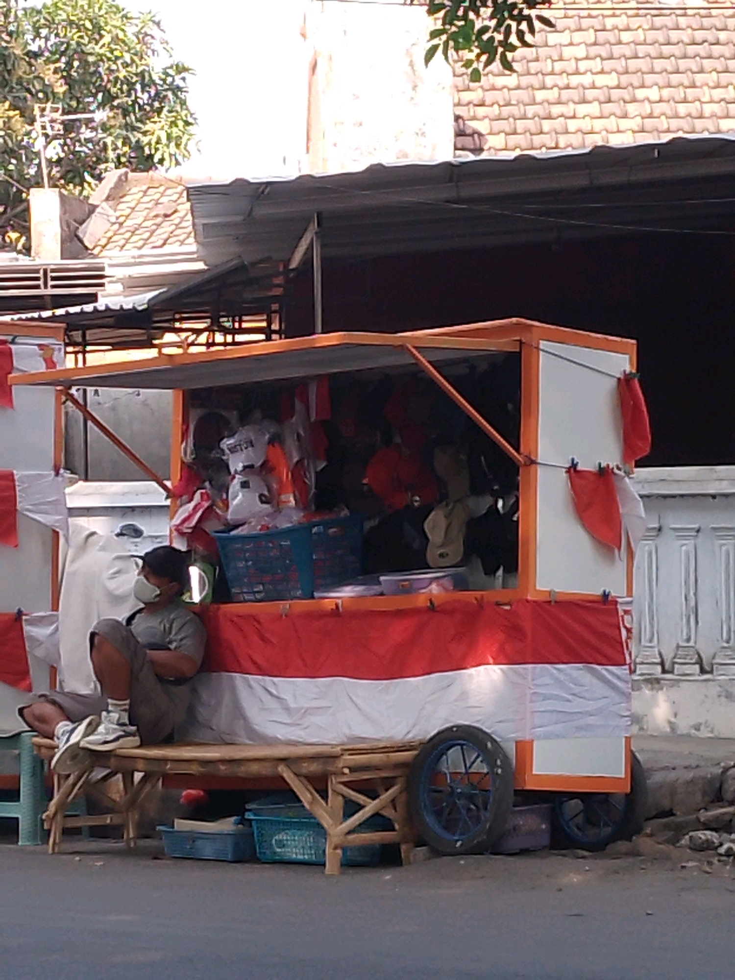 A person wearing a face mask and a grey t-shirt is sitting on a wooden bench in front of a stall. The stall is decorated with Indonesian flags. The person is wearing grey shorts, white socks, and white sneakers. The stall has an orange-trimmed roof and is covered with red and white fabric. Various items, including baskets and bags, are visible inside the stall. The stall is on wheels, suggesting it may be mobile. The background shows buildings and trees. The scene appears to be on a street in Kertosono, Indonesia.
