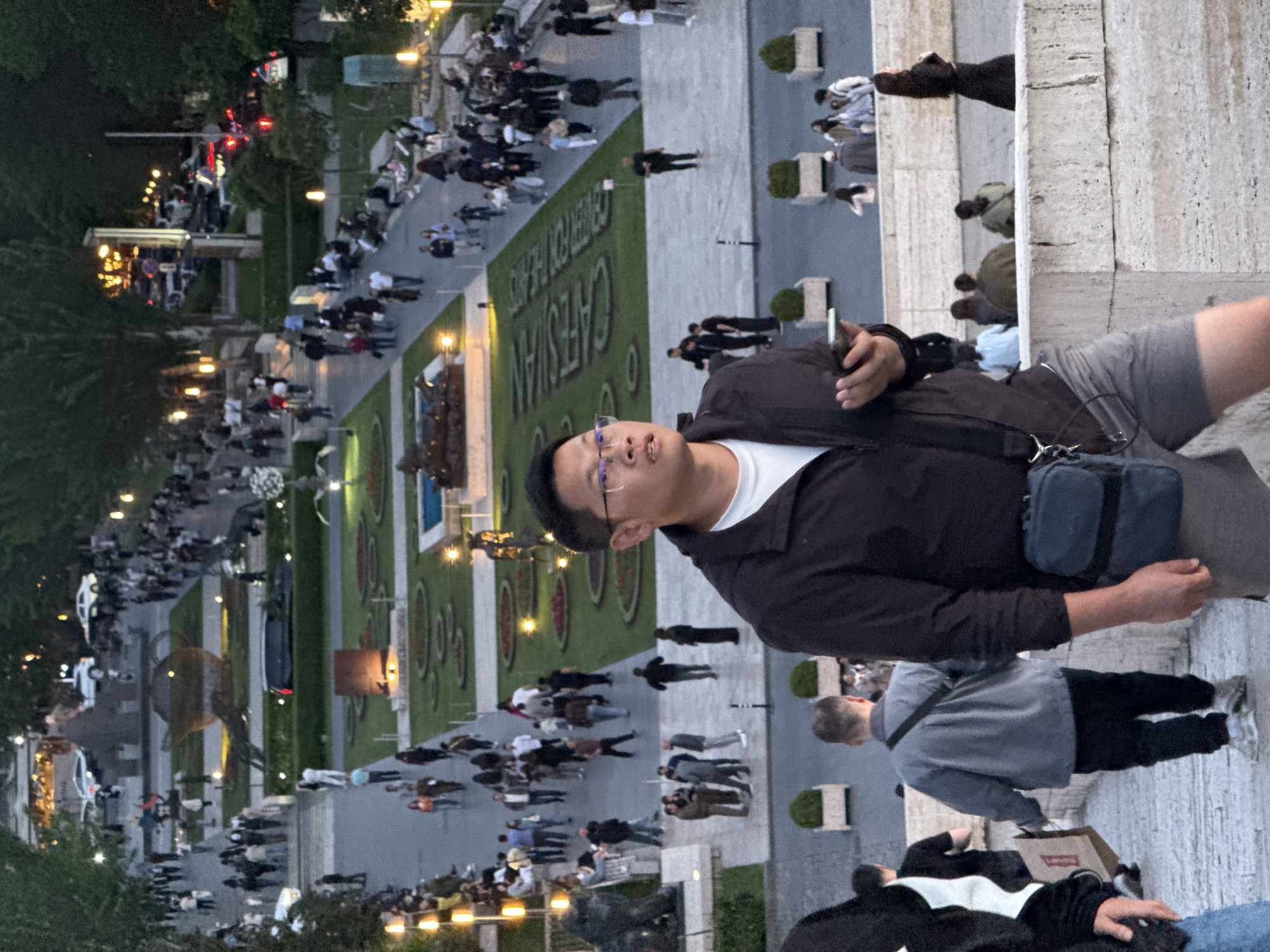 The image shows a street scene in Yerevan, Armenia, likely photographed from a high vantage point. The location appears to be near the Cascade complex, evident from the landscaping and layout. Many people are walking along the pedestrian areas, suggesting a busy evening. The man in the foreground is wearing glasses, a dark jacket, and light-colored shorts. He has a bag across his body and is holding a mobile phone. Another person wearing a grey shirt and black pants is standing next to him. There are lights lining the walkways, and vehicles can be seen in the distance.