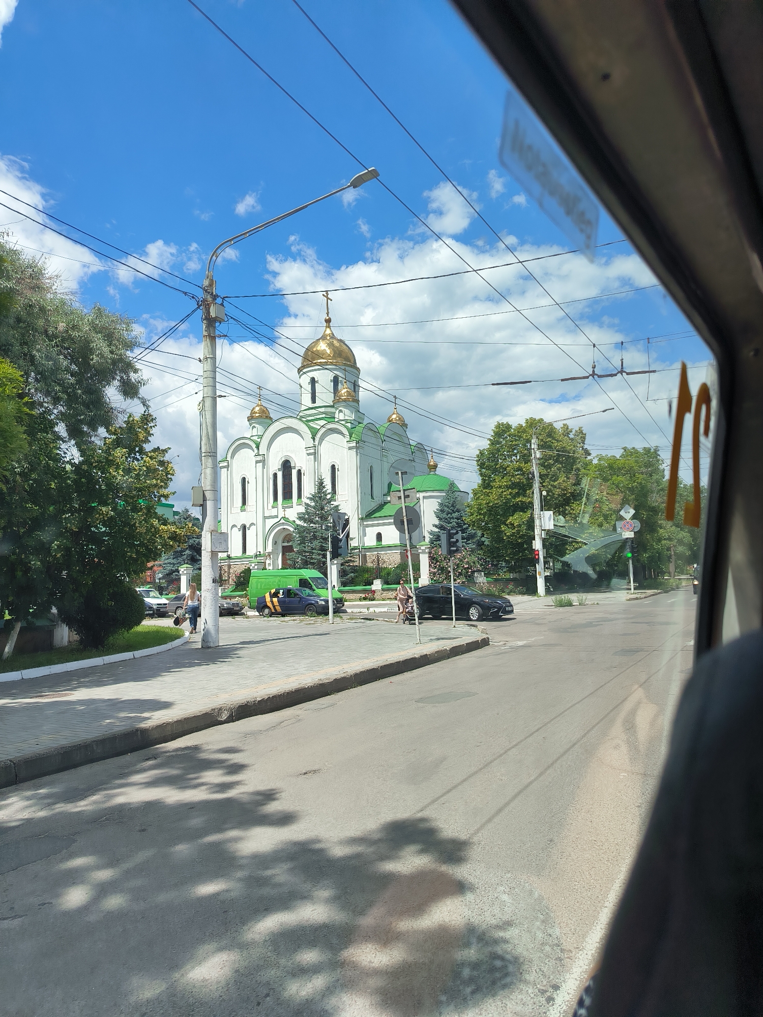 This is a medium-angle, full-frame shot taken from a moving vehicle, looking out a window. The scene captures a street in Tiraspol, Moldova, with a white Orthodox church featuring prominent golden domes and green trim dominating the background. A sidewalk with pedestrians and parked vehicles runs in front of the church. A traffic light is visible. The sky is blue with scattered white clouds, and overhead power lines crisscross the frame. The right side of the image shows the interior of the vehicle, including part of the window frame and a reflection of the outside scene.