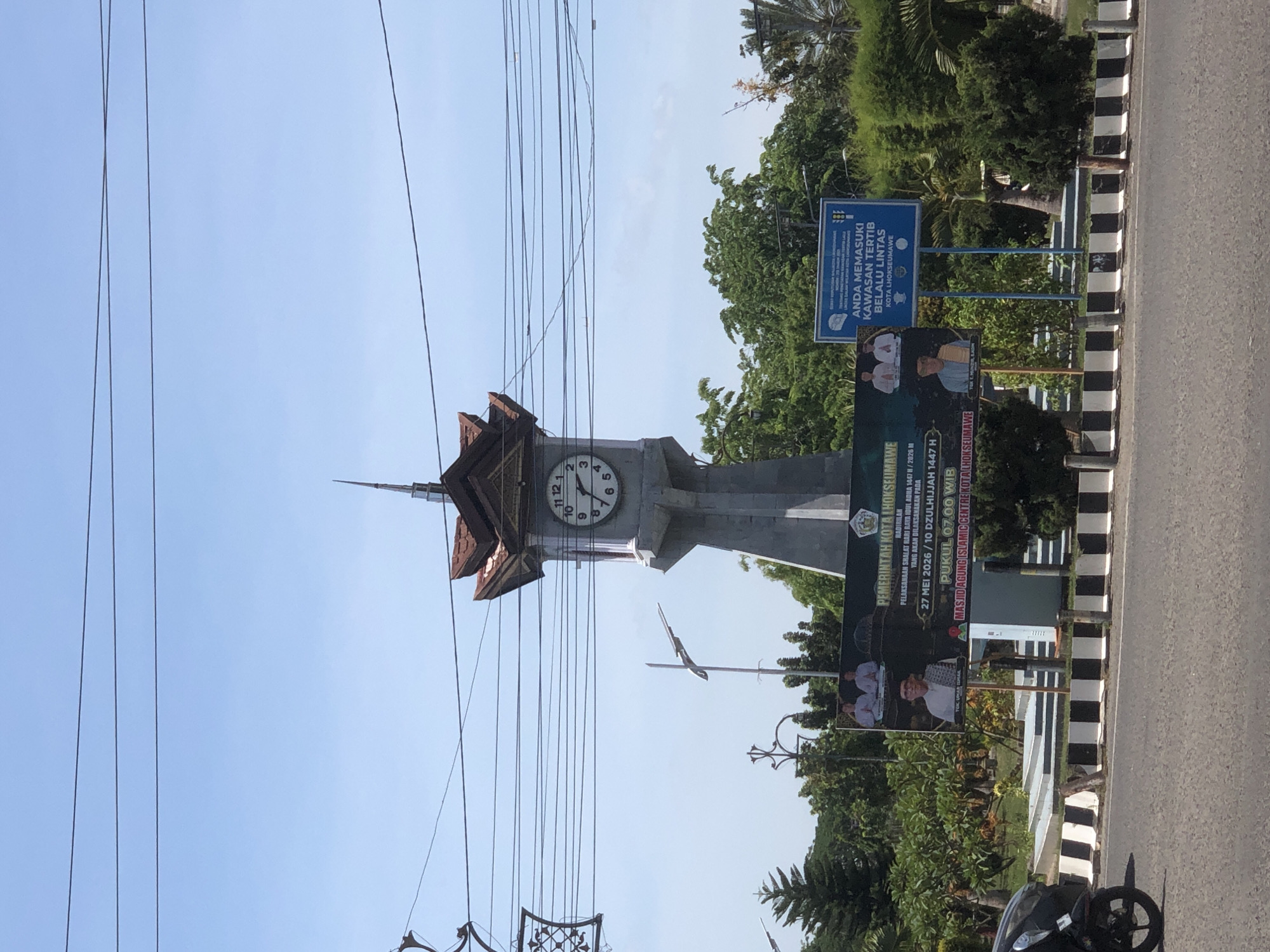 The image depicts a clock tower in Lhokseumawe, Indonesia. It's a light grey tower topped with a brown roof with a clock face visible. Power lines crisscross the foreground. A sign advertising the upcoming Eid al-Adha prayer at the Islamic Centre Mosque is prominent. Another sign reads, "Anda Memasuki Kawasan Tertib Berlalu Lintas Kota Lhokseumawe," indicating a traffic-regulated zone. A motorcycle is partially visible on the right. Vegetation and trees are in the background. The sky is blue.