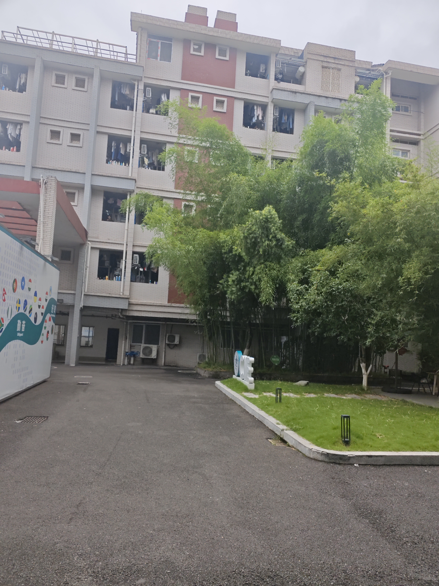 The image captures an outdoor scene in the Chongqing High-tech Zone, China, on a cloudy day. In the foreground, a paved asphalt area leads to a grassy area bordered by a concrete curb. Two small, black, rectangular outdoor lamps are positioned on the grass. To the left, a white rectangular mural with a teal wavy design and various colorful circular emblems is visible.
Behind the mural and the paved area, a multi-story building with a light brick facade and grey accents stands. Several windows are open, with laundry hanging out to dry. Air conditioning units are visible on some of the building's exterior walls. A thick stand of bamboo and other green trees forms a dense background, partially obscuring the building. The overall impression is of a residential or administrative building complex with some landscaping. No people are clearly visible in the image.
